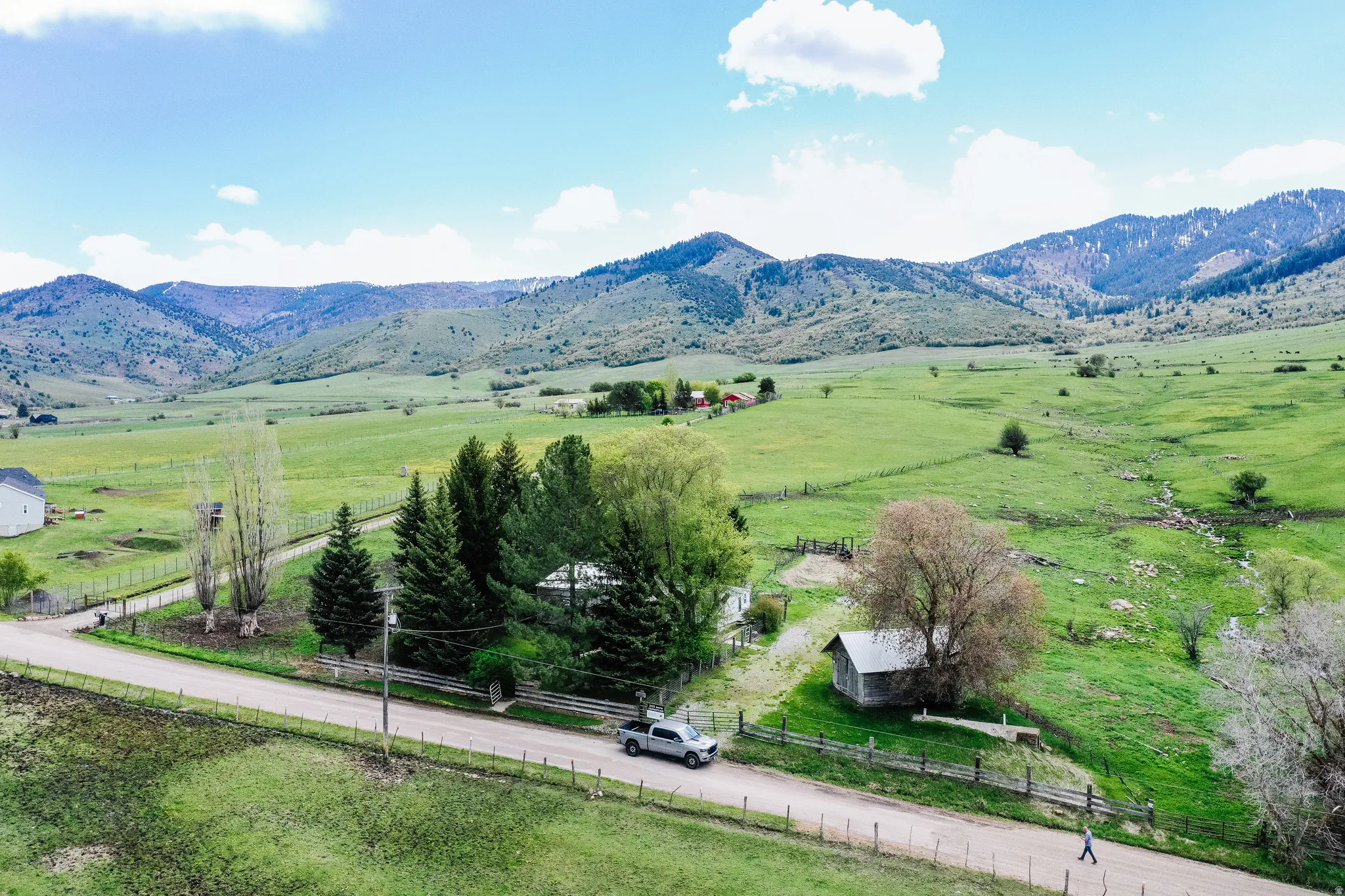 Expansive green landscape with distant mountains, featuring a gravel road, mature trees, and fencing
