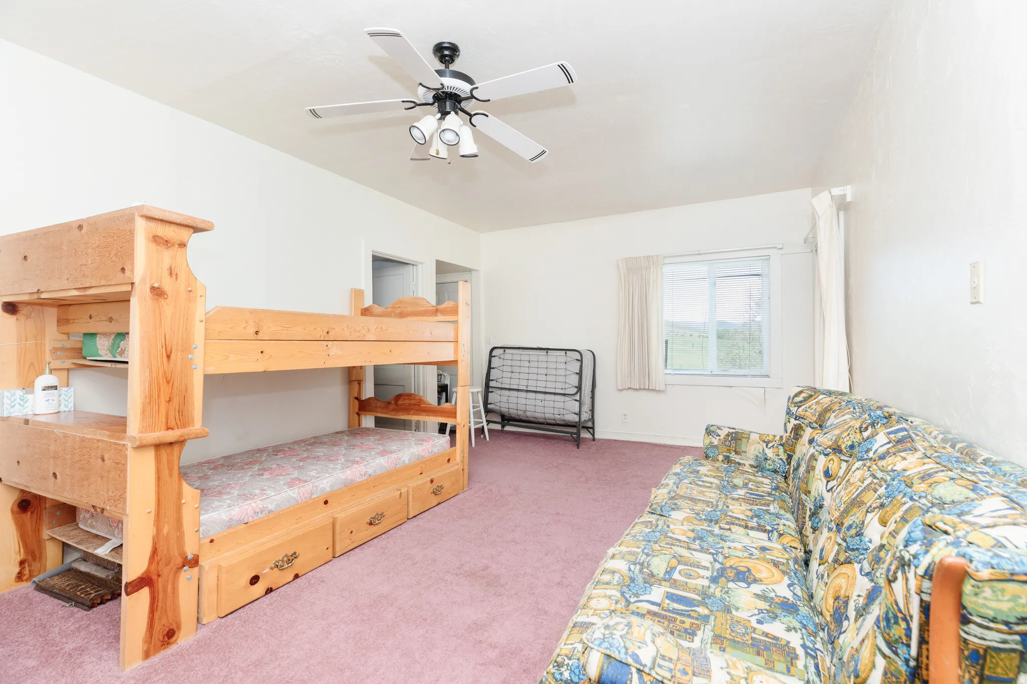 Spacious bedroom suite featuring built-in wooden bunk beds with integrated storage drawers, ceiling fan with lighting, plush carpeting throughout, and abundant natural light from large window with white curtain panels