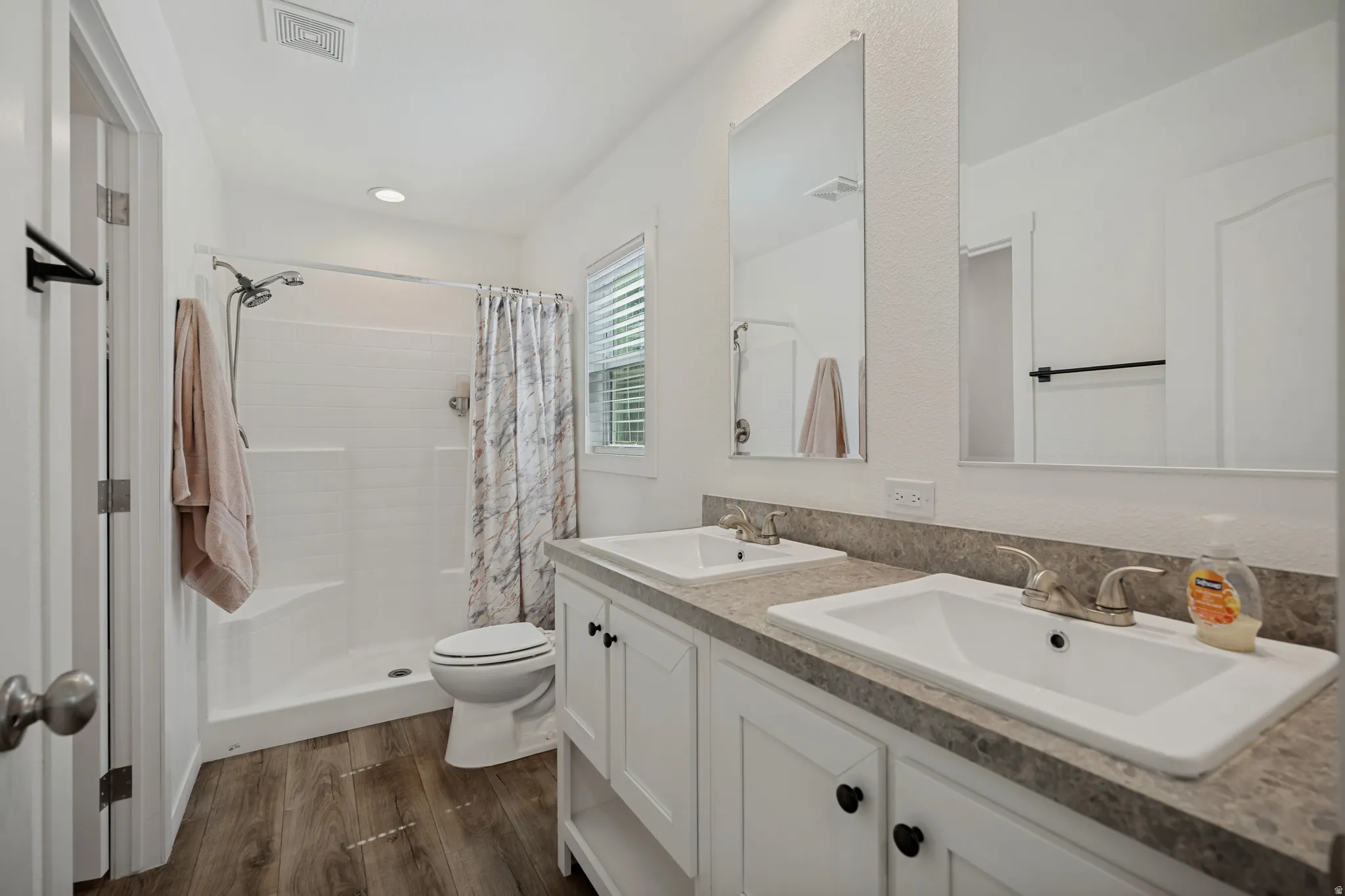 Dual vanity with integrated sinks, wood-finish flooring, white cabinetry with dark hardware, a shower-tub combination, and a window with blinds