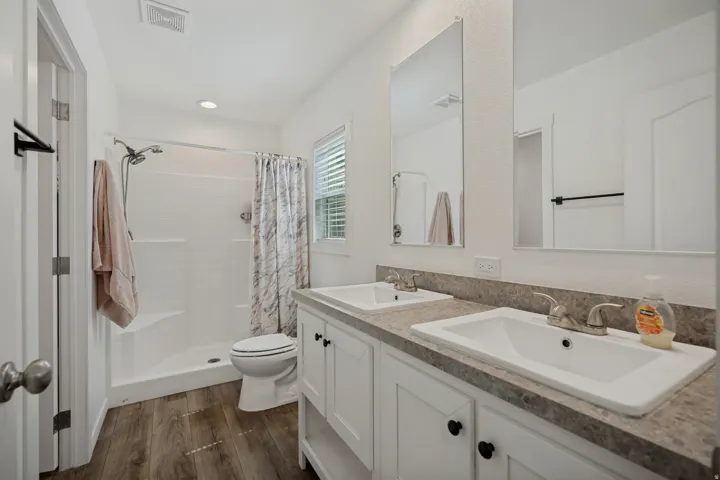 Dual vanity with integrated sinks, wood-finish flooring, white cabinetry with dark hardware, a shower-tub combination, and a window with blinds