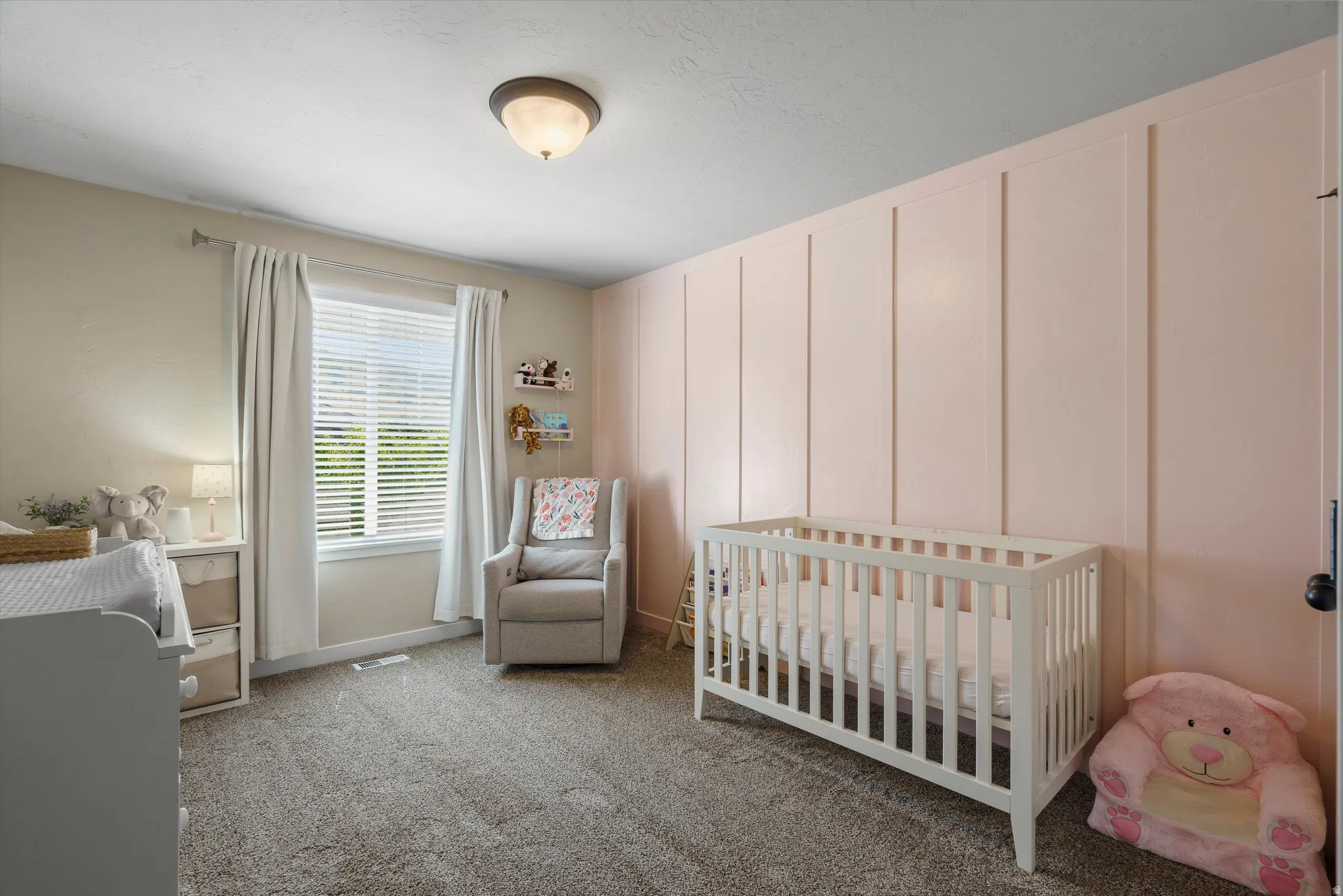 Wall with vertical paneling detail, wall-to-wall carpeting, window with blinds and curtains, ceiling-mounted light fixture, and a built-in changing station