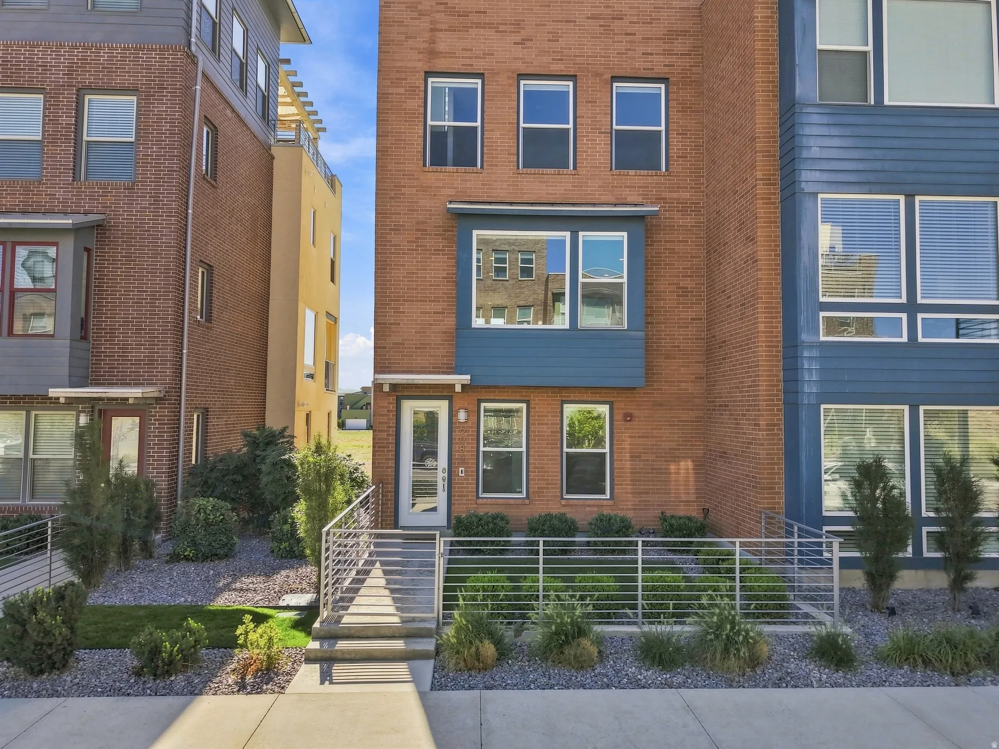 Brick exterior with modern blue accent siding, multiple windows across three levels, concrete entrance with metal railings, and landscaped front yard
