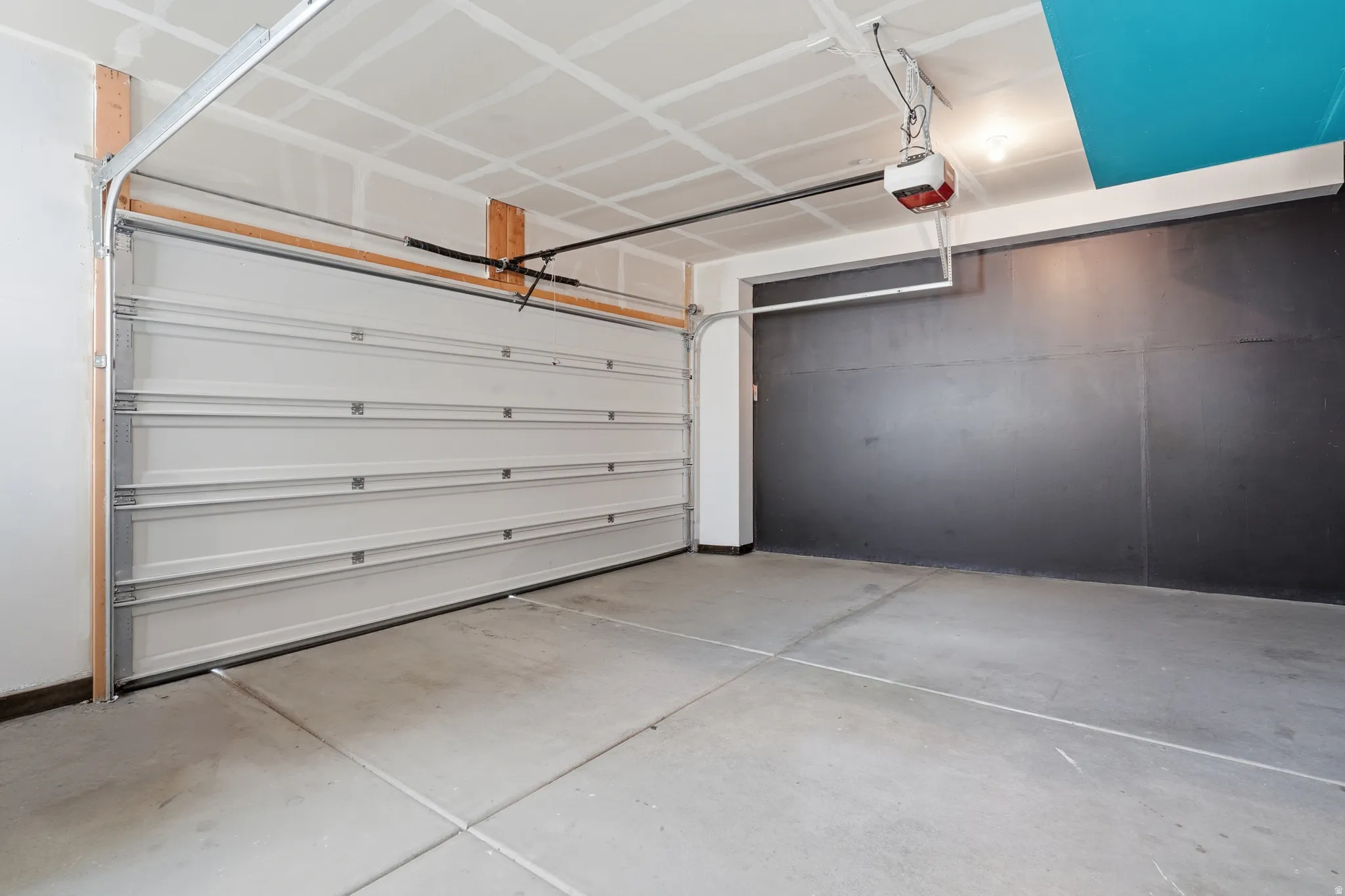 Concrete flooring with expansion joints, white paneled garage door, exposed ceiling joists, and a wall-mounted garage door opener