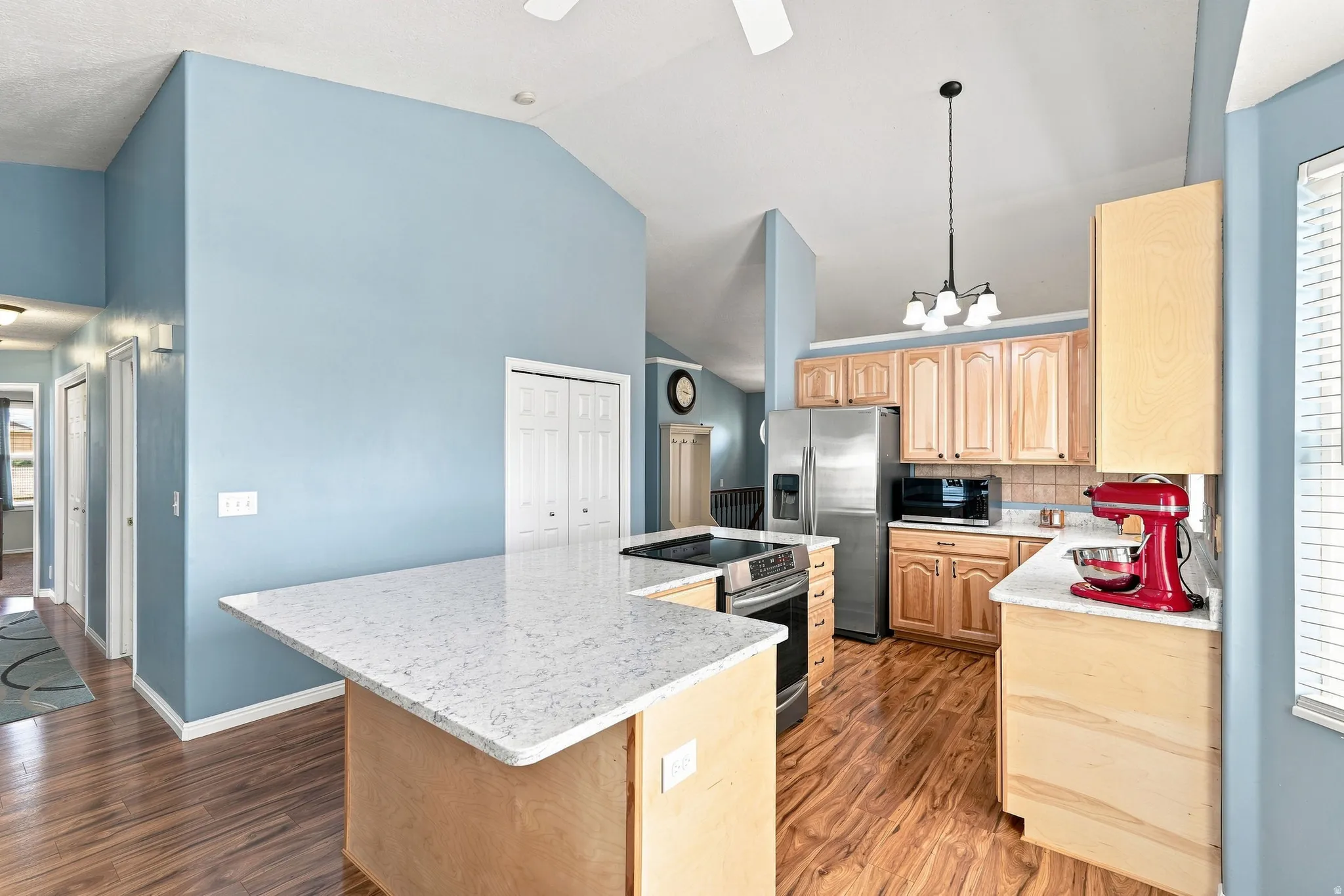 Kitchen featuring vaulted ceilings, wood-finish flooring, light wood cabinetry, white stone-finish countertops, and stainless steel appliances