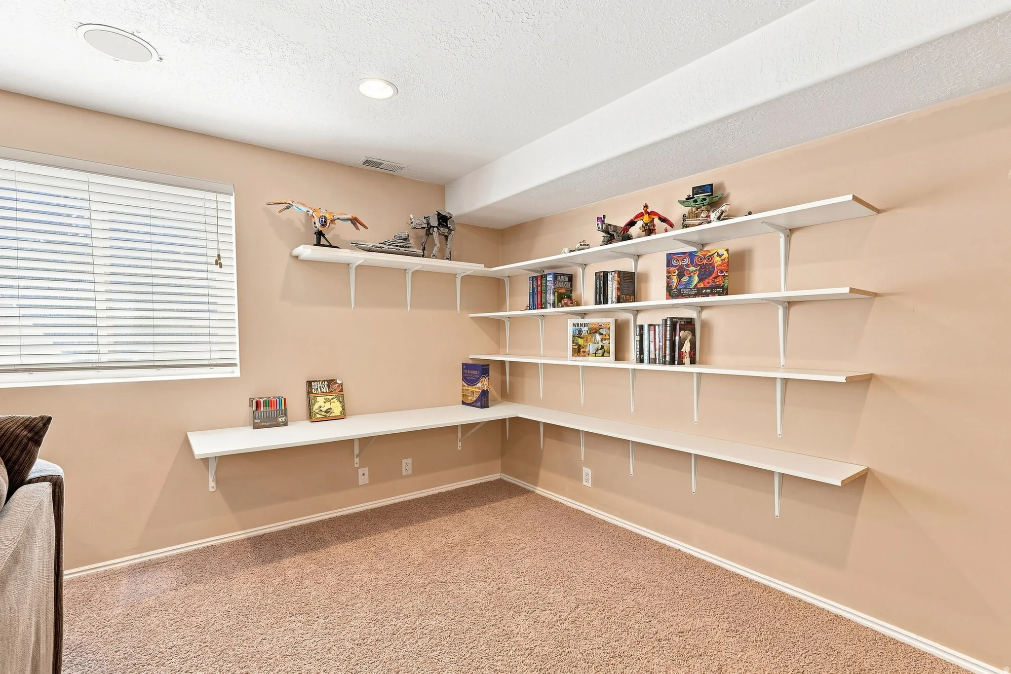 Wall-mounted shelving system, recessed ceiling lighting, textured carpeting, window with horizontal blinds, integrated ceiling speaker