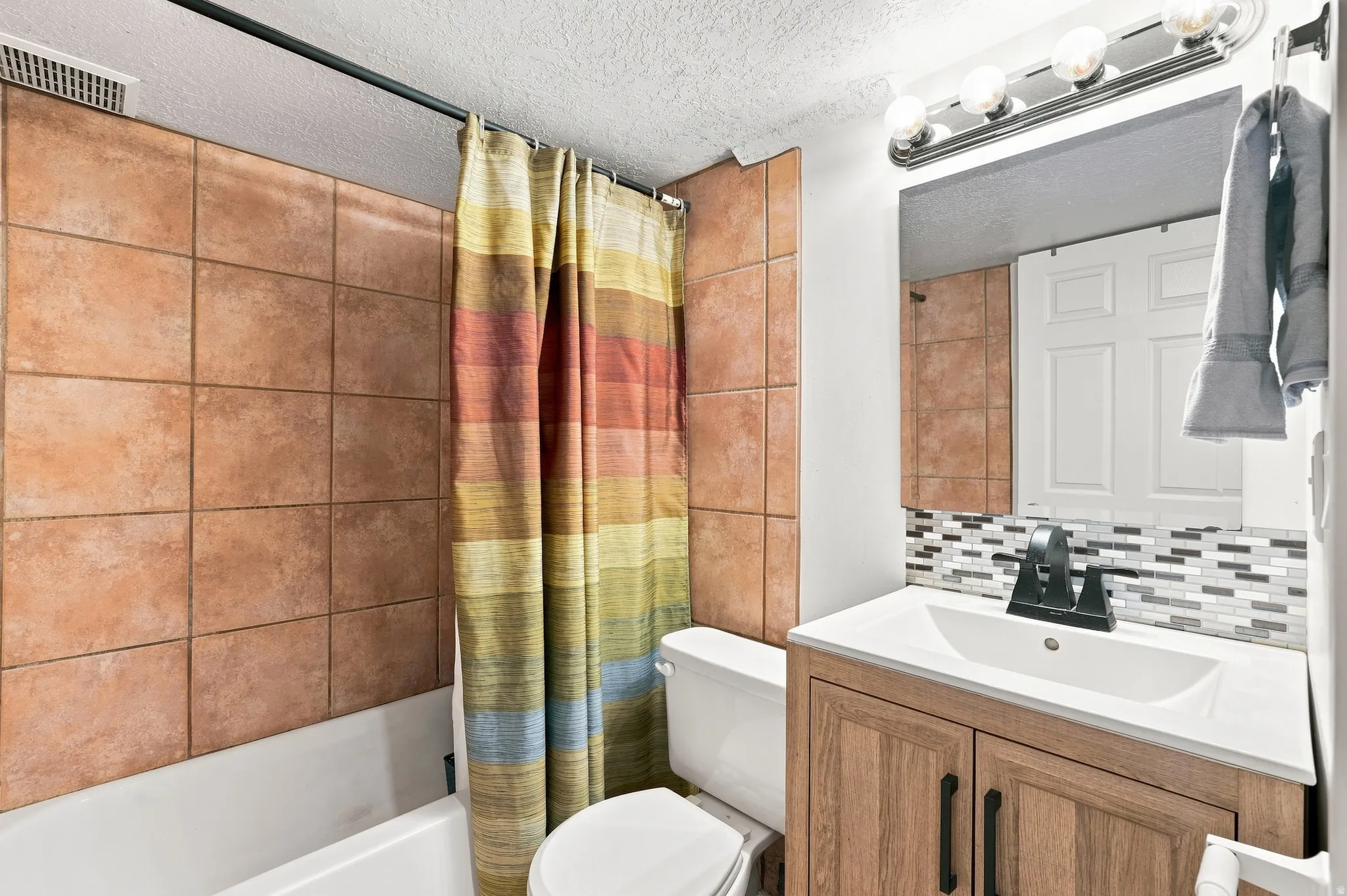 Bathroom featuring a built-in bathtub with tiled surround, a vanity with wood-finish cabinetry, an integrated sink, a mosaic tile backsplash, and a wall-mounted mirror with overhead lighting