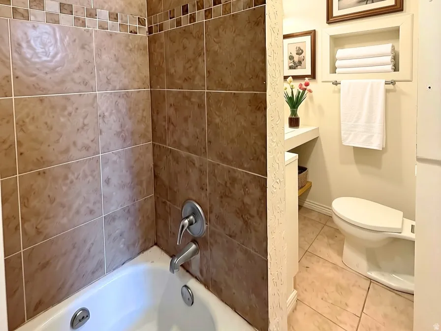 Bathtub with tile surround, built-in recessed shelving, toilet, and tile flooring