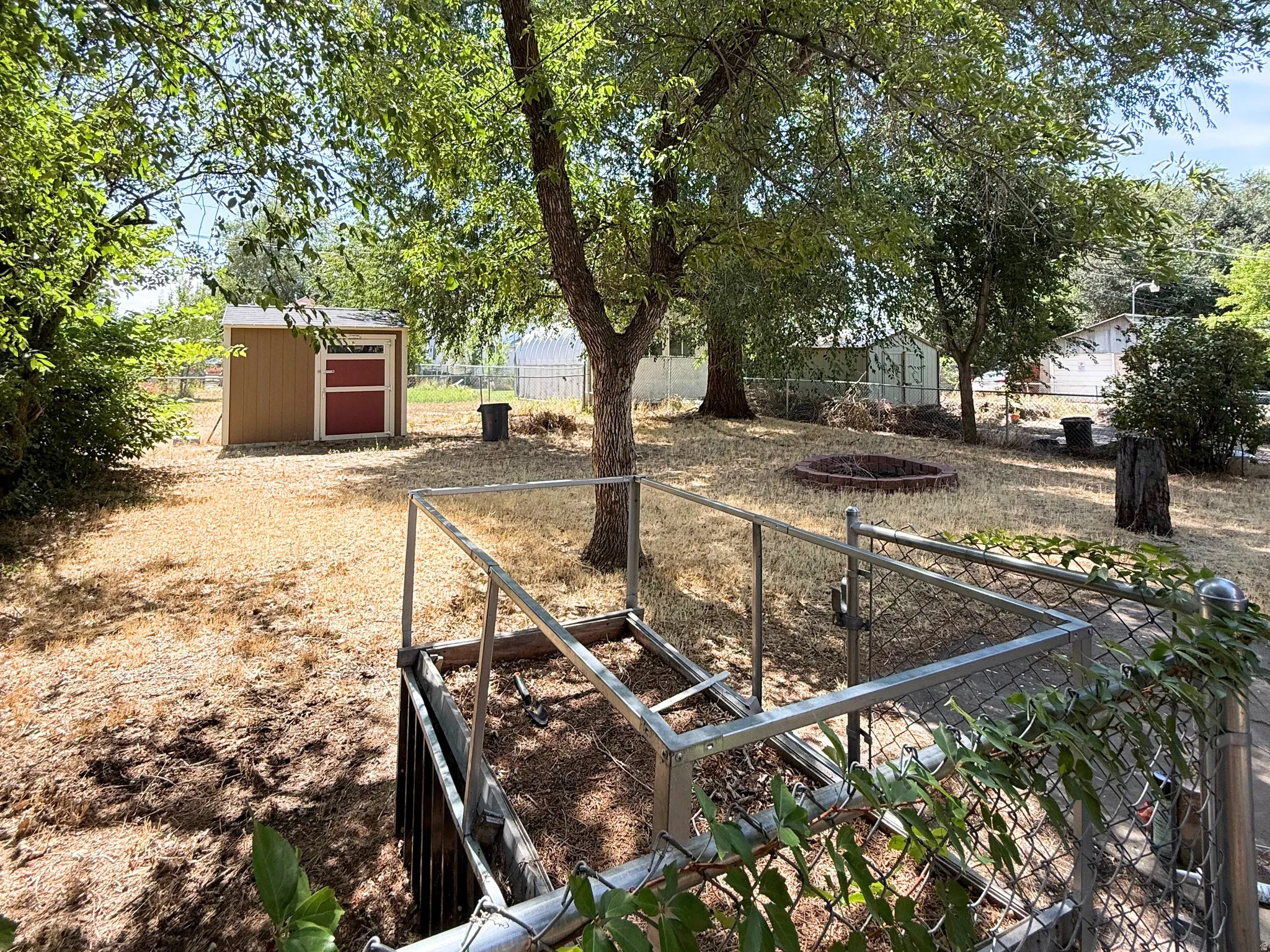 Expansive backyard featuring mature trees, a detached storage shed, a chain-link fence, and a built-in fire pit