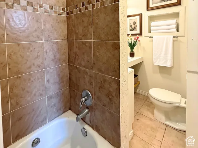 Bathtub with tile surround, built-in recessed shelving, toilet, and tile flooring