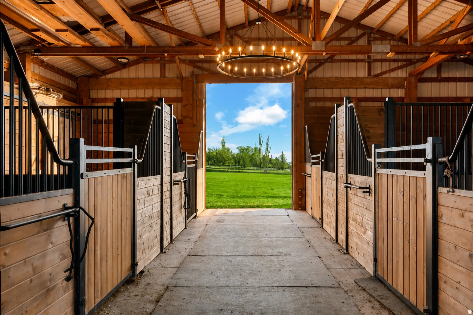 Barn interior with wood beam construction, multiple stalls featuring wood paneling and black metal accents, and a central concrete aisle