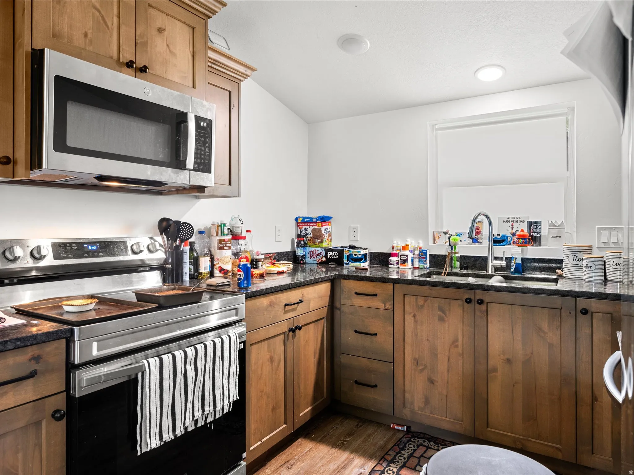 Wood-finish cabinetry with dark hardware, dark countertops, stainless steel microwave, stainless steel range, and undermount sink with a gooseneck faucet