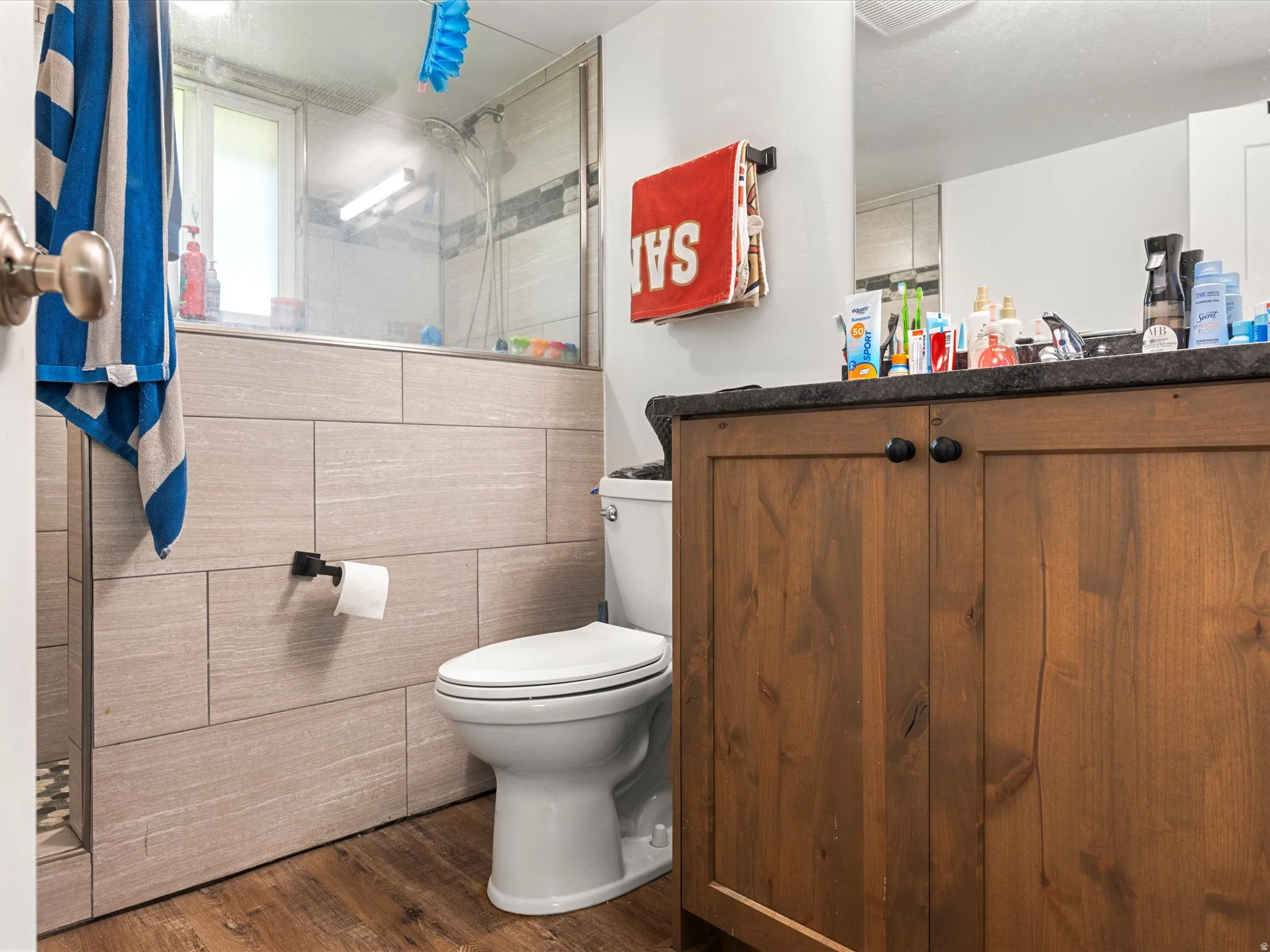 Wood-finish flooring, wood vanity with black hardware, dark countertop, white toilet, and a walk-in shower with glass enclosure