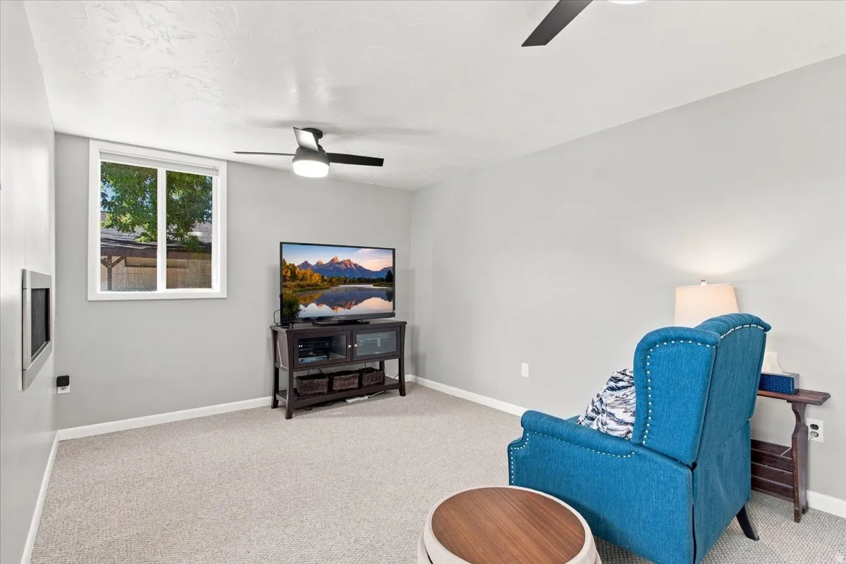 Recessed wall fireplace, light gray wall paint, window with white trim, light-colored carpet flooring, and ceiling fan