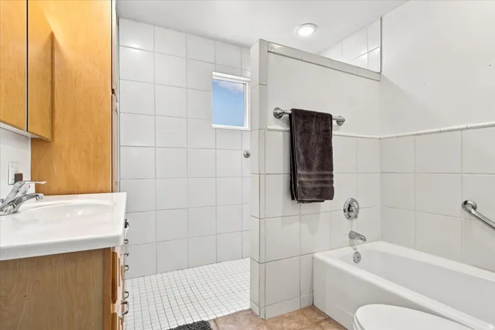 Full bathroom featuring a white tile shower with a small window, a separate bathtub, a vanity with wood-finish cabinetry, and a white countertop with an integrated sink