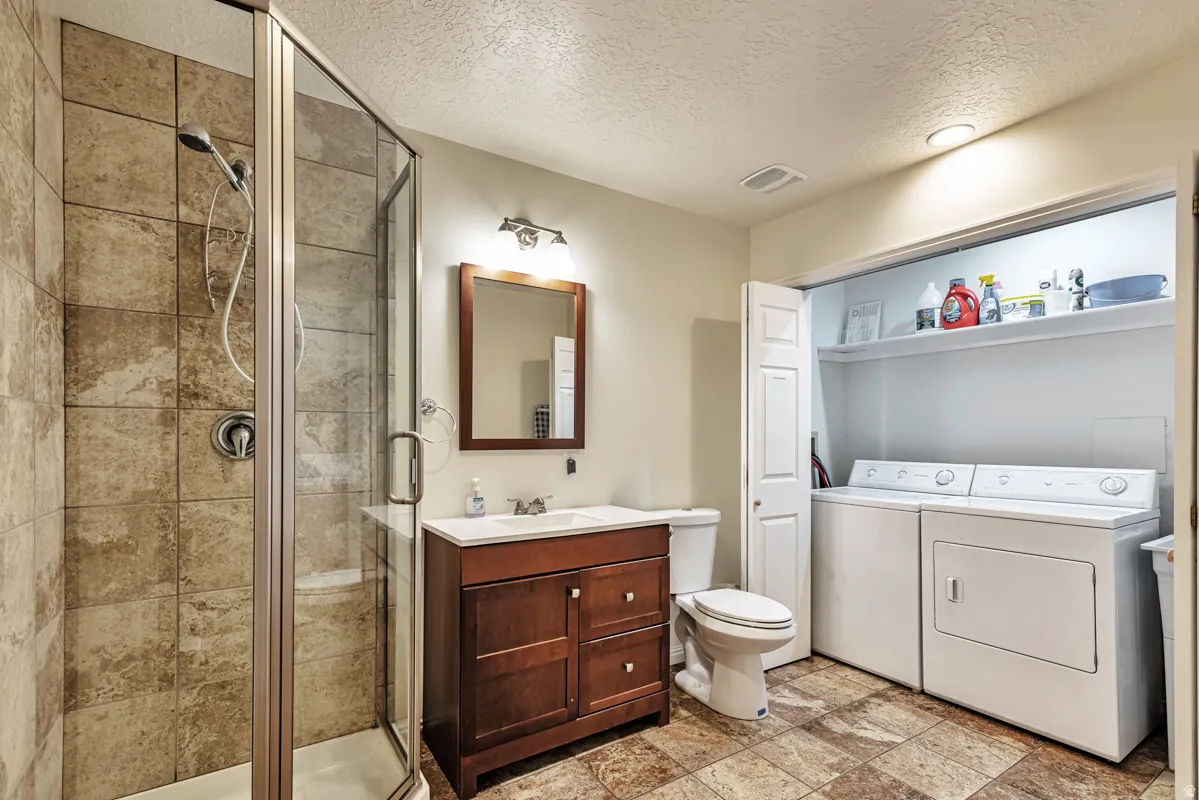 Glass-enclosed shower with tiled walls, wood-finish vanity with countertop, wall-mounted mirror, and built-in laundry area with shelving