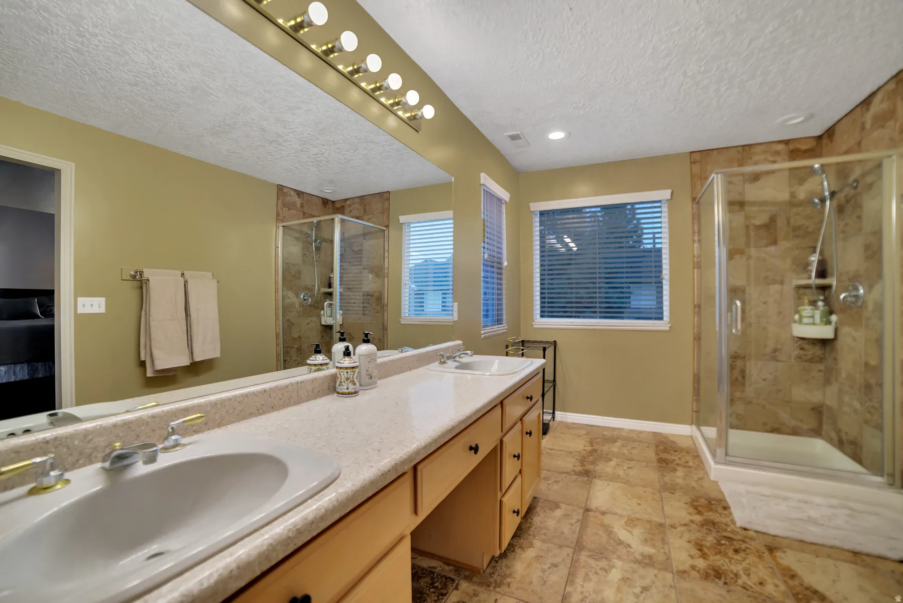 Dual vanity with integrated sinks, full-width mirror with overhead lighting, glass-enclosed shower, and tiled flooring