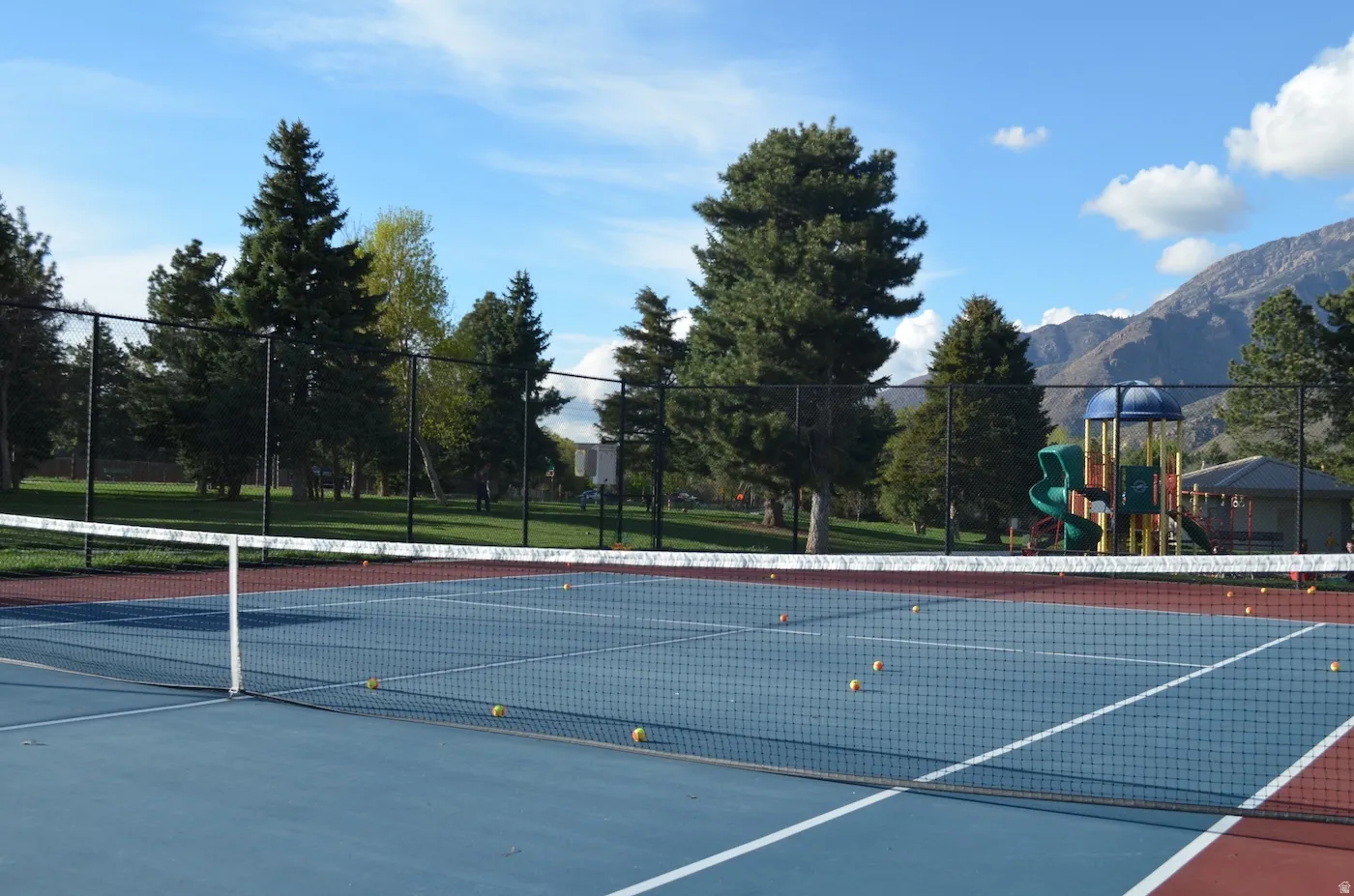 Blue and red tennis court with a black chain-link fence, surrounded by mature trees, and a mountain backdrop