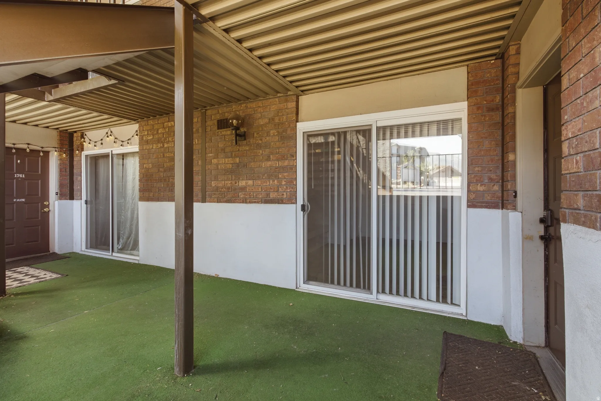 Covered patio area with artificial turf flooring, brick facade, and sliding glass door access