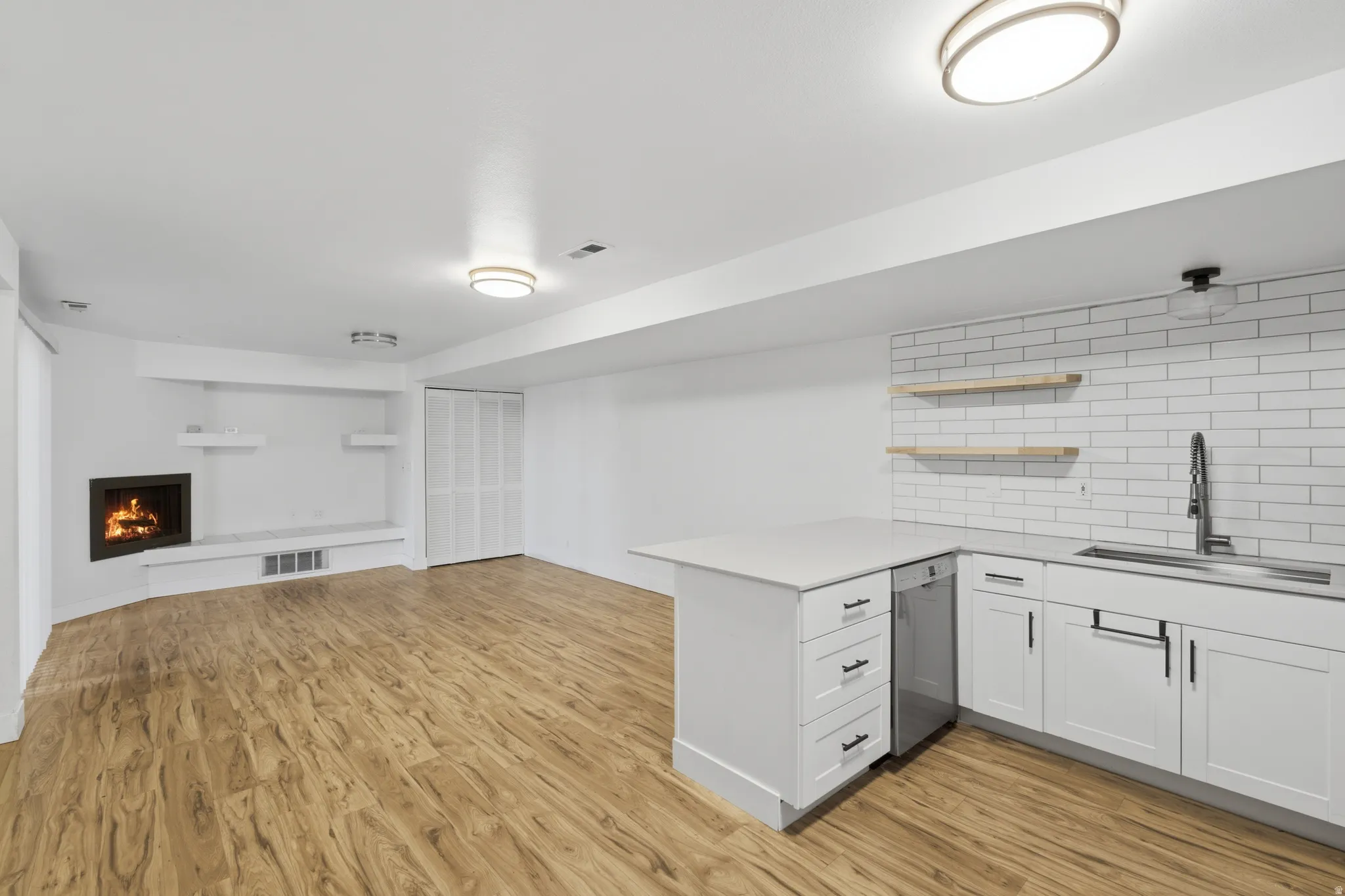 Wood-finish flooring throughout, featuring a built-in fireplace with hearth, white bi-fold closet doors, a kitchen area with white subway tile backsplash, and open wood shelving