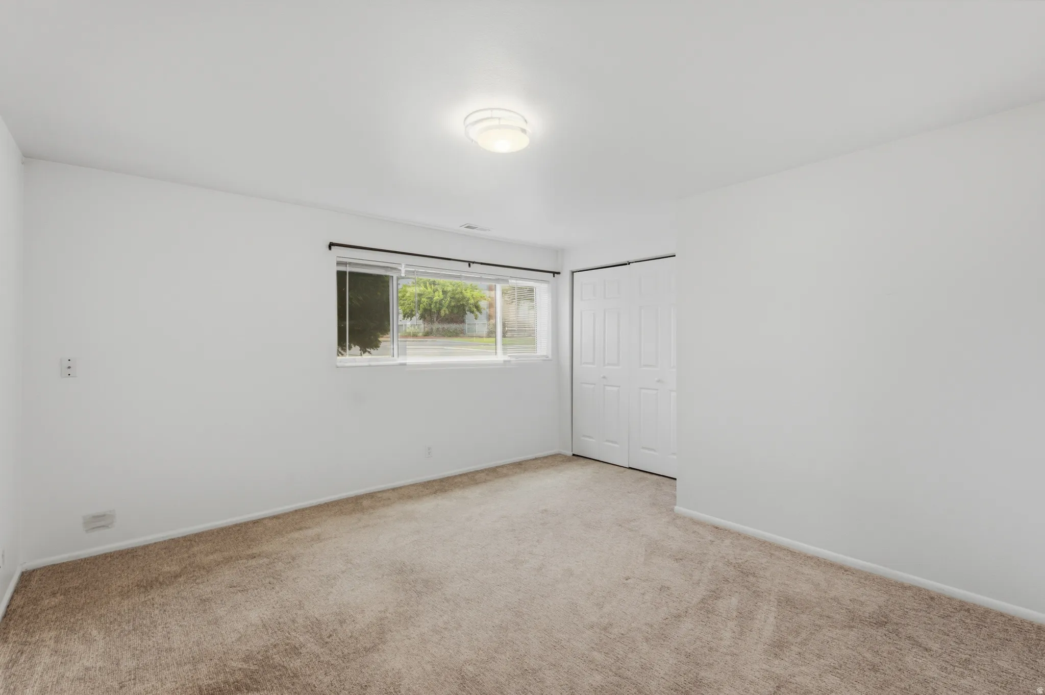 Carpeted flooring, white walls, built-in bi-fold closet, ceiling-mounted light fixture, and a window with exterior visibility
