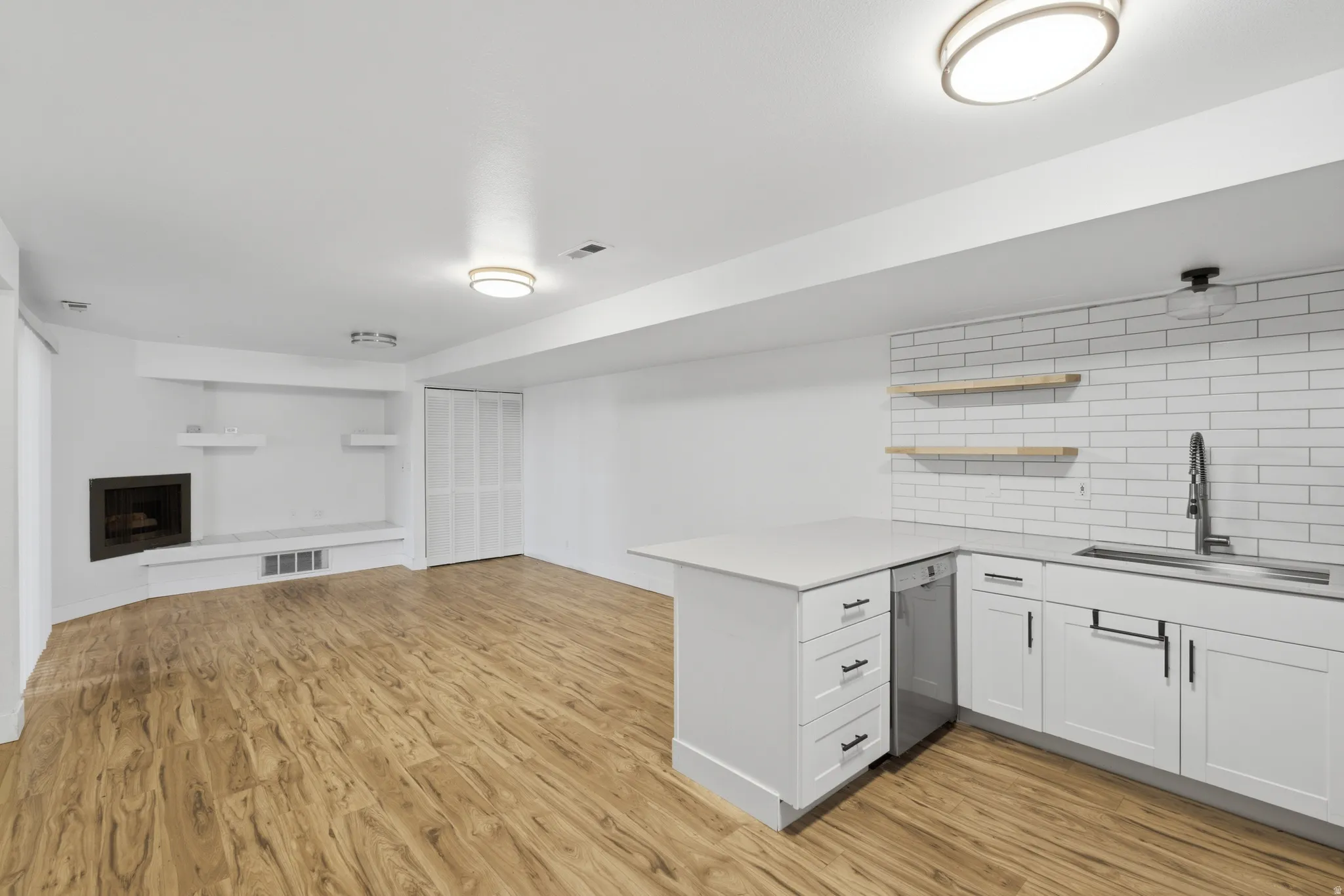 Wood-finish flooring throughout, featuring a fireplace with built-in shelving, white bi-fold closet doors, and a kitchenette with white shaker cabinetry, white subway tile backsplash, and floating wood shelves