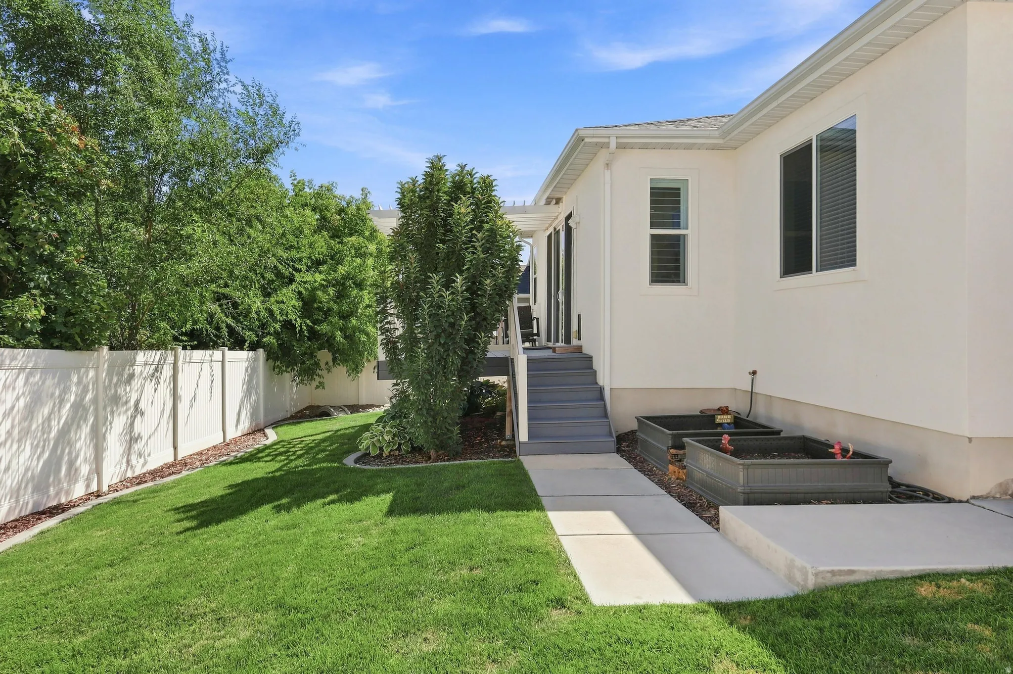 Manicured lawn with mature perimeter trees, light-toned stucco exterior, raised garden beds, and a white privacy fence