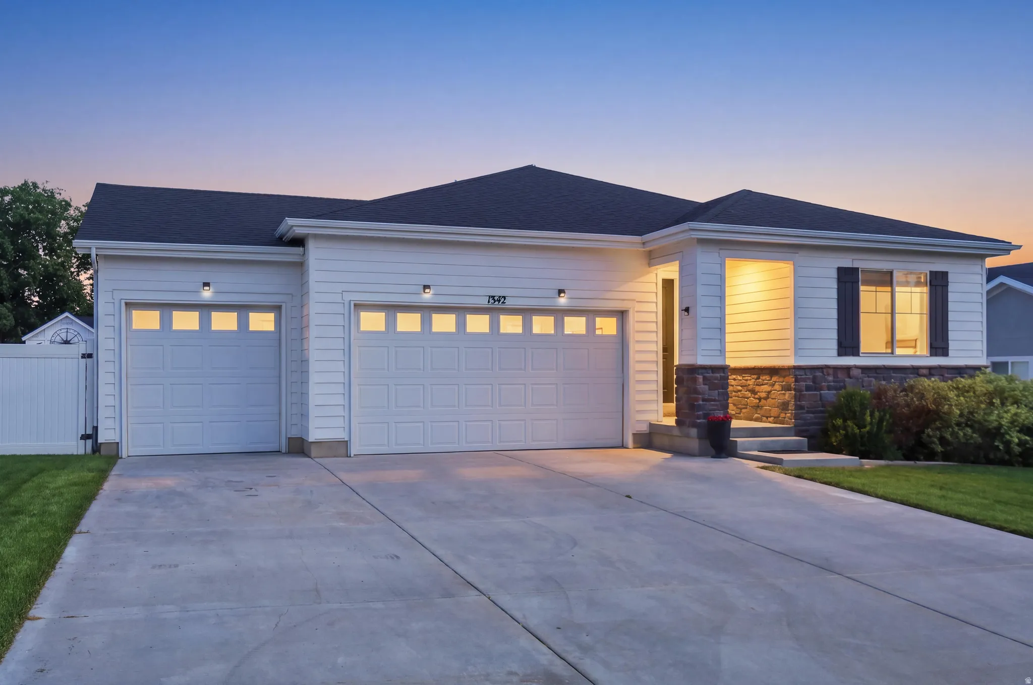 White siding exterior with dark shingles, stone accent facade, two-car garage with paneling, concrete driveway, and covered entryway