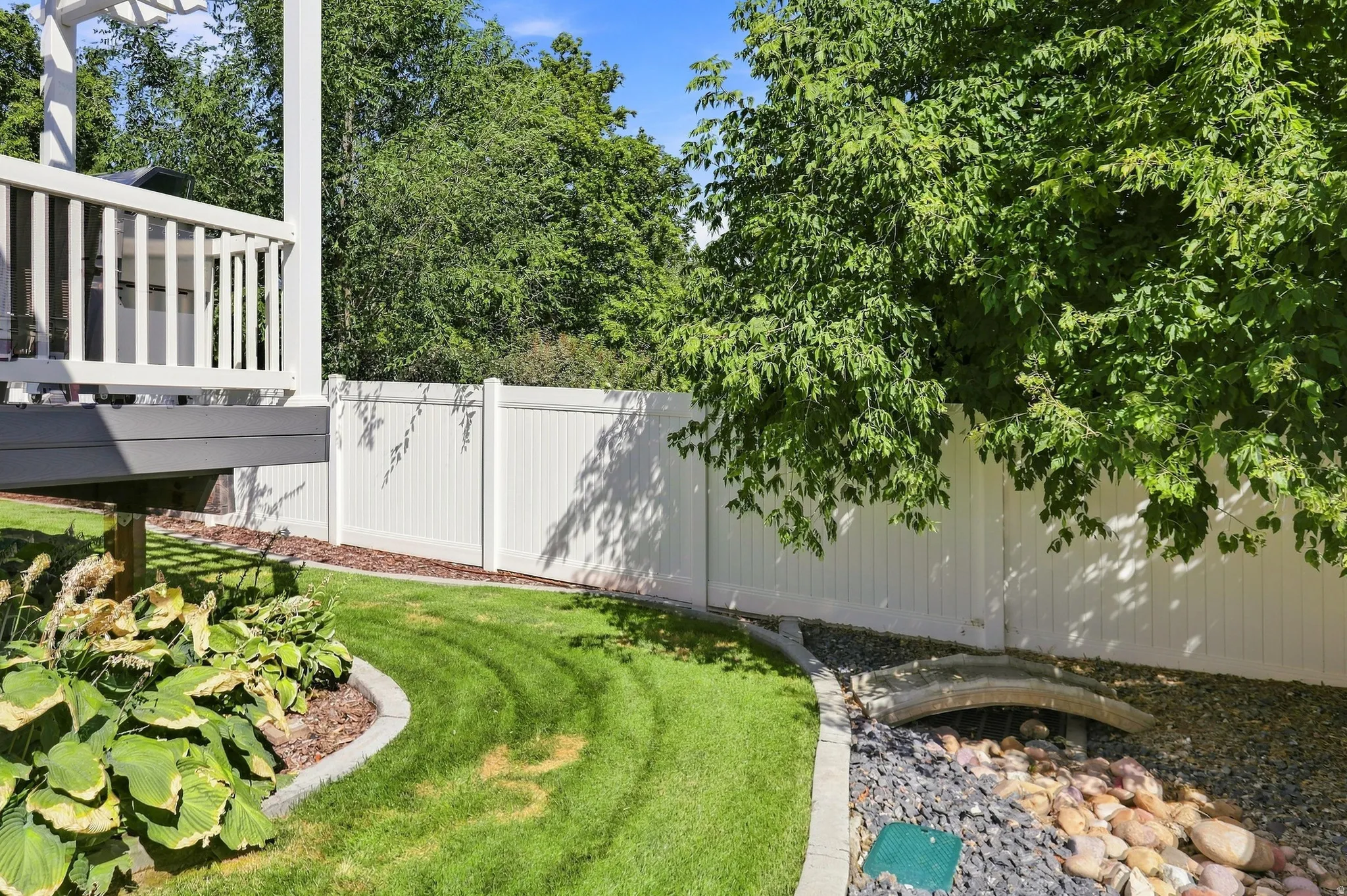 Backyard featuring a white privacy fence, mature trees, and a decorative rock bed with a bridge