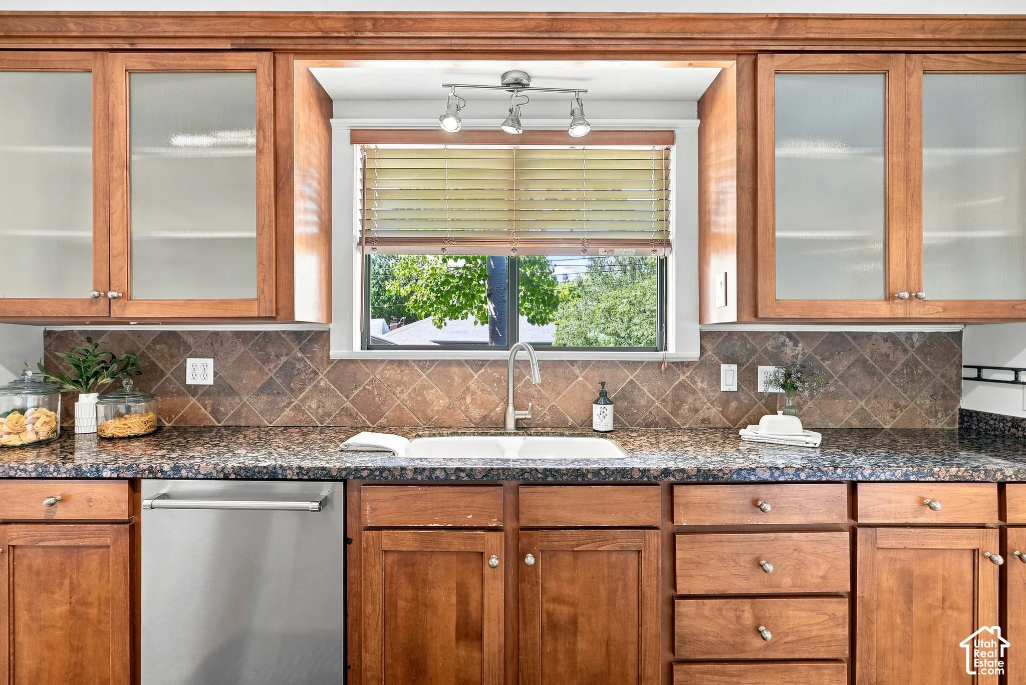 Wood cabinetry with frosted glass inserts, speckled countertops, diamond-patterned backsplash, undermount sink with gooseneck faucet, and stainless steel dishwasher