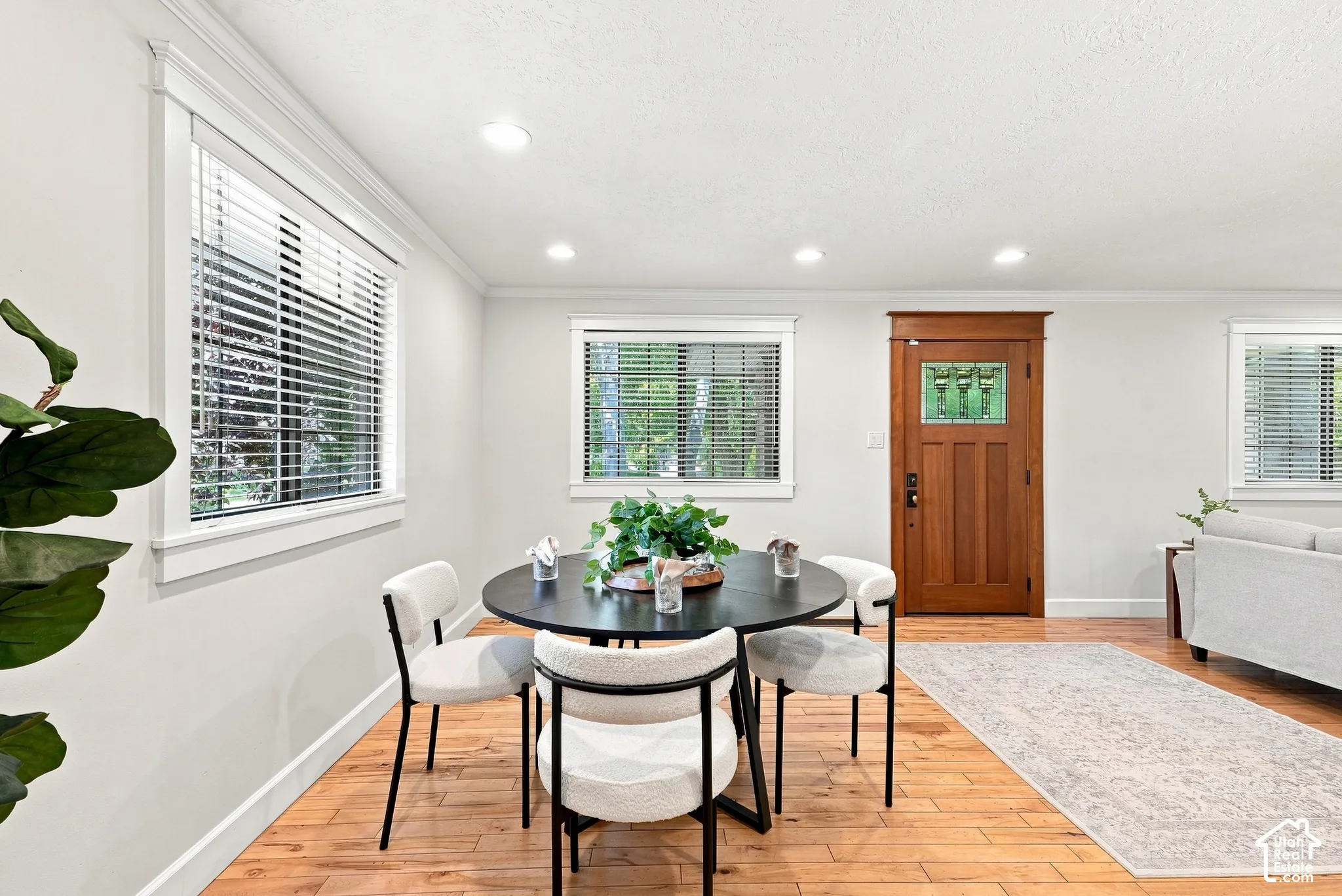 Bright dining area featuring a round black table with wood-finish flooring, recessed ceiling lighting, and multiple windows with plantation shutters