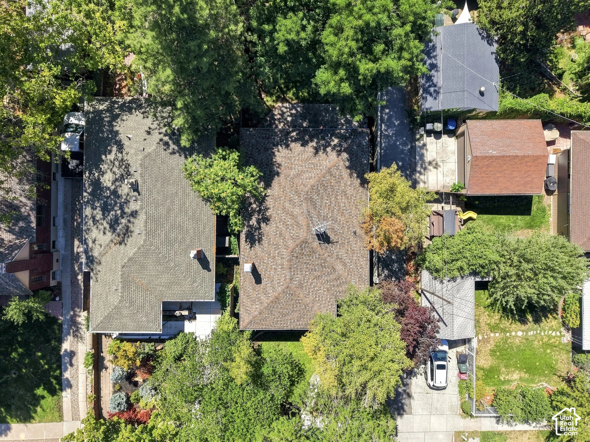 Aerial view showcasing mature landscaping with established trees throughout the property, multiple roof lines with dimensional asphalt shingles, brick exterior construction, and a manicured lawn with defined hardscaping elements