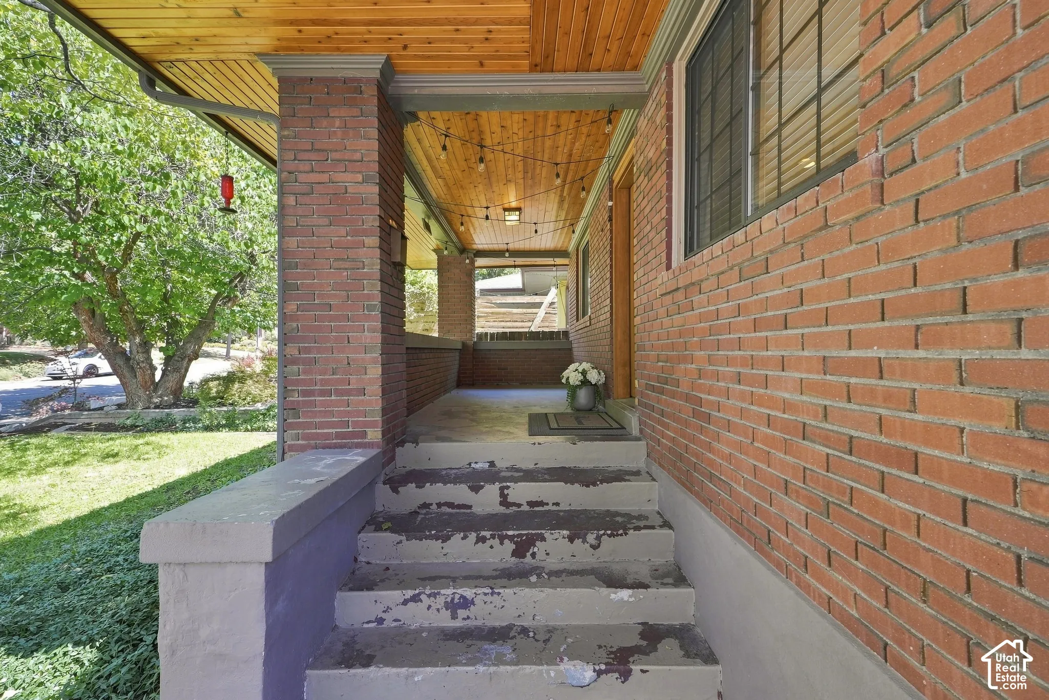 Covered entry breezeway featuring exposed wood-beam ceiling, brick exterior walls, concrete stepping stones, and mature landscaping with established trees visible from the approach