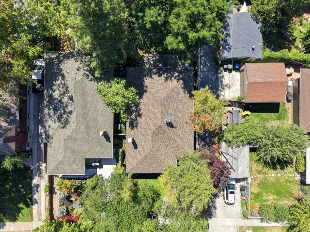 Aerial view showcasing mature landscaping with established trees throughout the property, multiple roof lines with dimensional asphalt shingles, brick exterior construction, and a manicured lawn with defined hardscaping elements