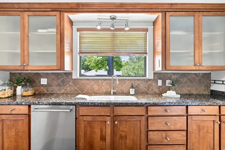 Wood cabinetry with frosted glass inserts, speckled countertops, diamond-patterned backsplash, undermount sink with gooseneck faucet, and stainless steel dishwasher
