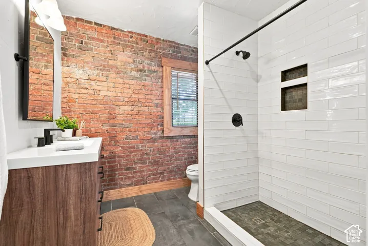 Modern primary bathroom featuring exposed brick accent wall, white subway tile shower surround with frameless glass enclosure, floating wood-finish vanity with integrated sink, matte black fixtures, and dark tile flooring