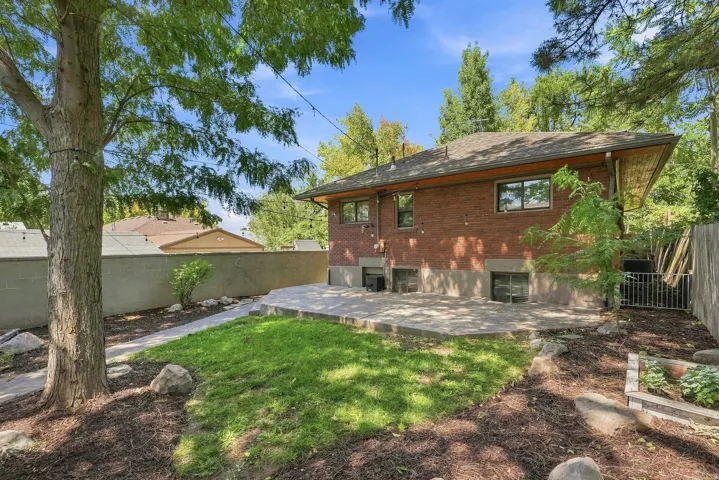 Brick Colonial residence with a spacious backyard featuring mature shade trees, a manicured lawn, mulched landscaping beds, and a rear patio area