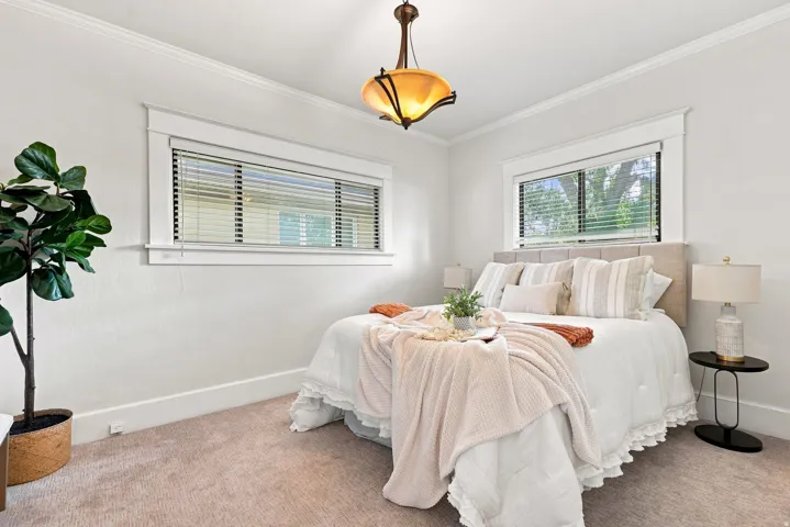 Bright, light-filled bedroom showcasing two generous windows with horizontal blinds, neutral wall finishes, and warm wood-finish flooring accented with neutral area carpeting
