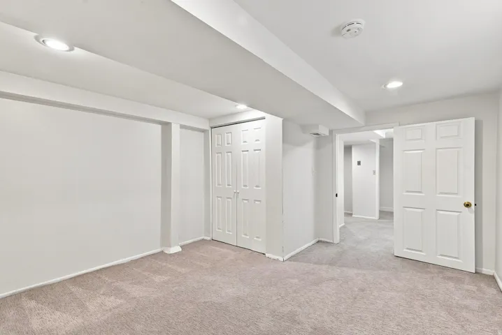 Spacious finished basement featuring neutral carpet flooring, recessed ceiling lighting, multiple white six-panel closet doors, and an open floor plan with structural support columns