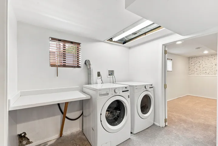 Modern laundry room featuring side-by-side front-load washing machines, white cabinetry with countertop workspace, skylights providing natural illumination, and neutral concrete flooring
