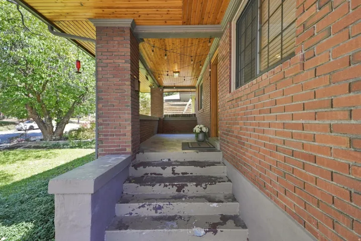 Covered entry breezeway featuring exposed wood-beam ceiling, brick exterior walls, concrete stepping stones, and mature landscaping with established trees visible from the approach