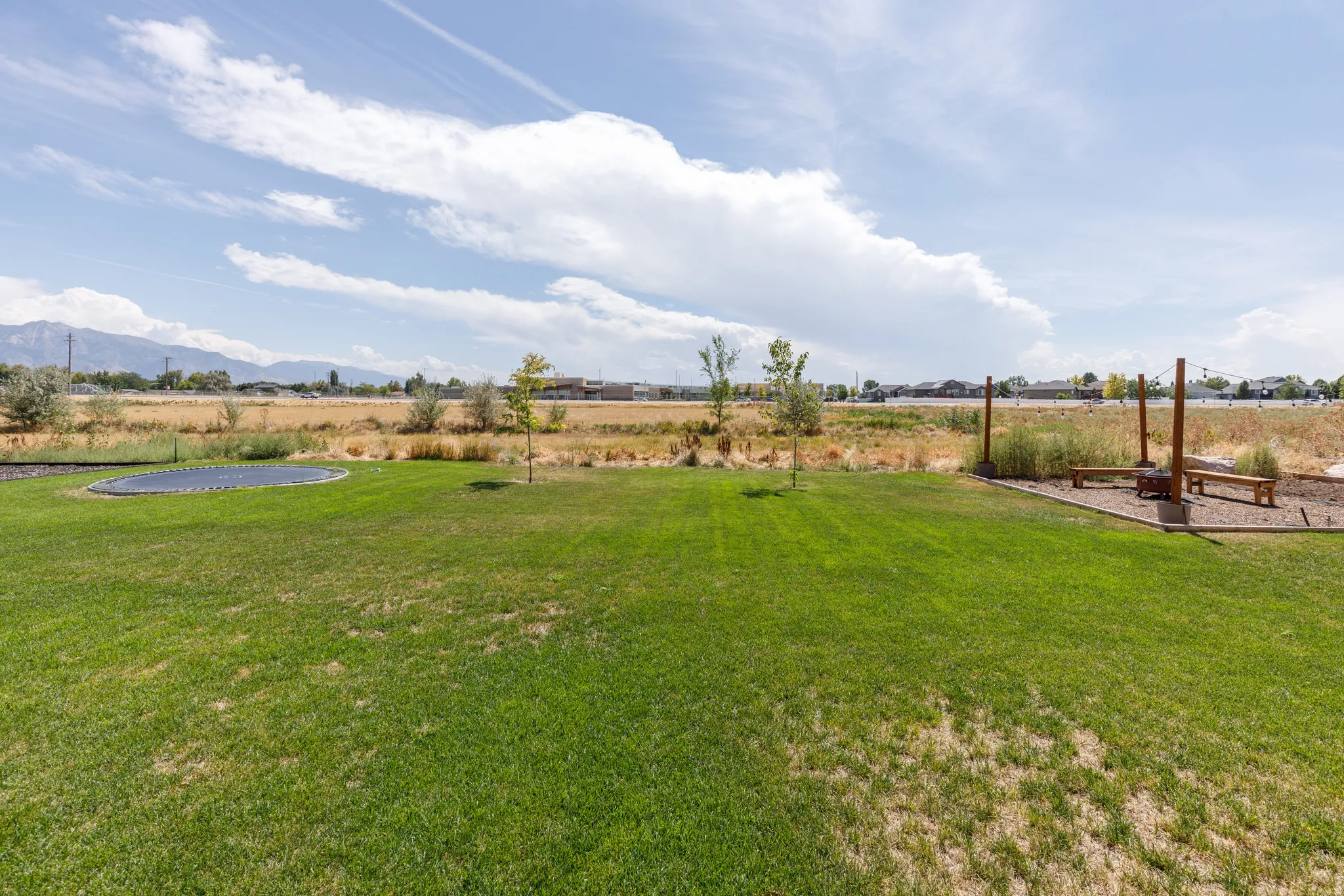 Expansive lawn area with a built-in trampoline, mature trees, and a gravel-covered entertainment space featuring permanent wooden benches and posts