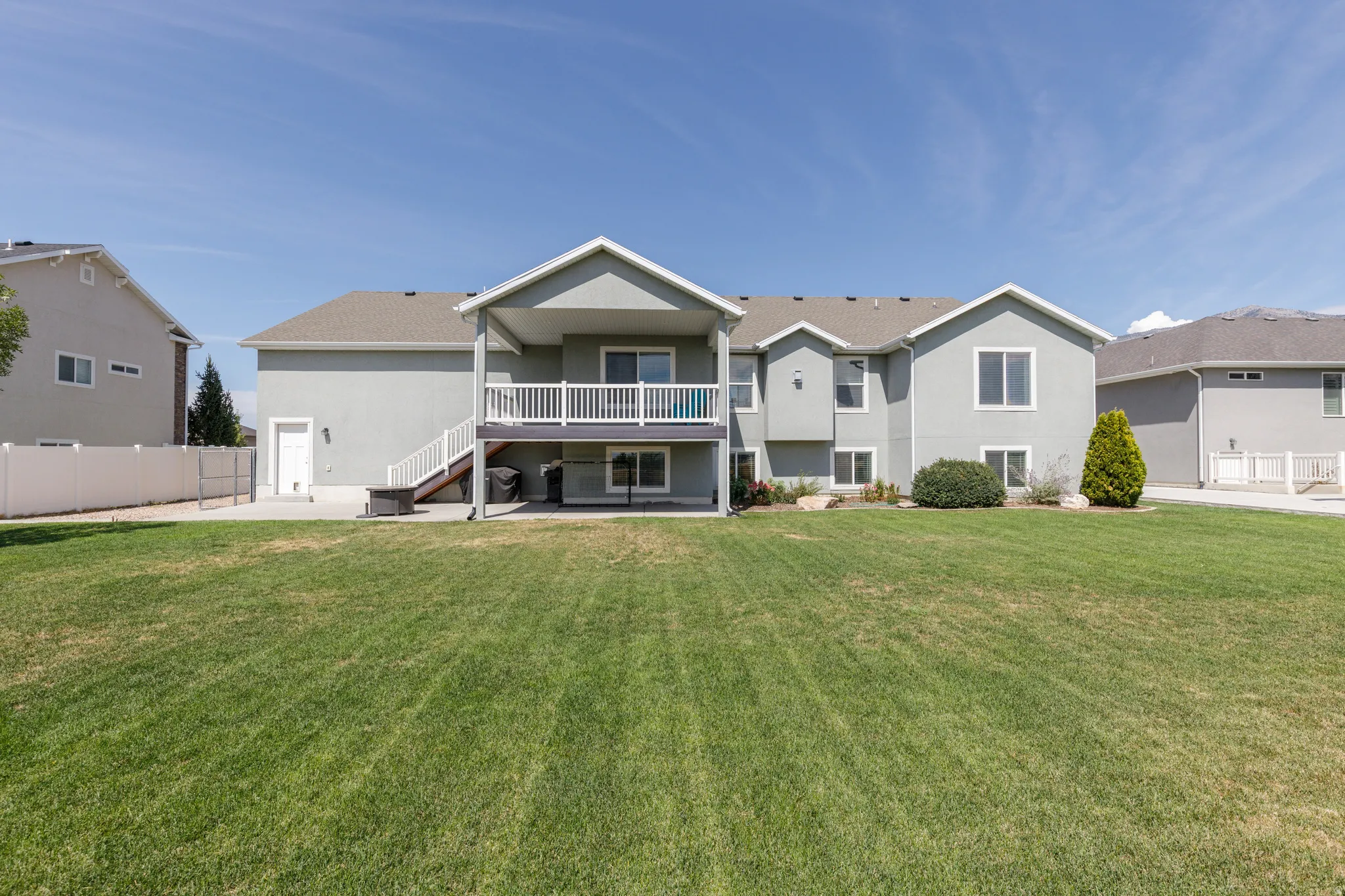 Expansive rear yard featuring a manicured lawn, stucco exterior, upper-level deck with white railings, and a concrete patio