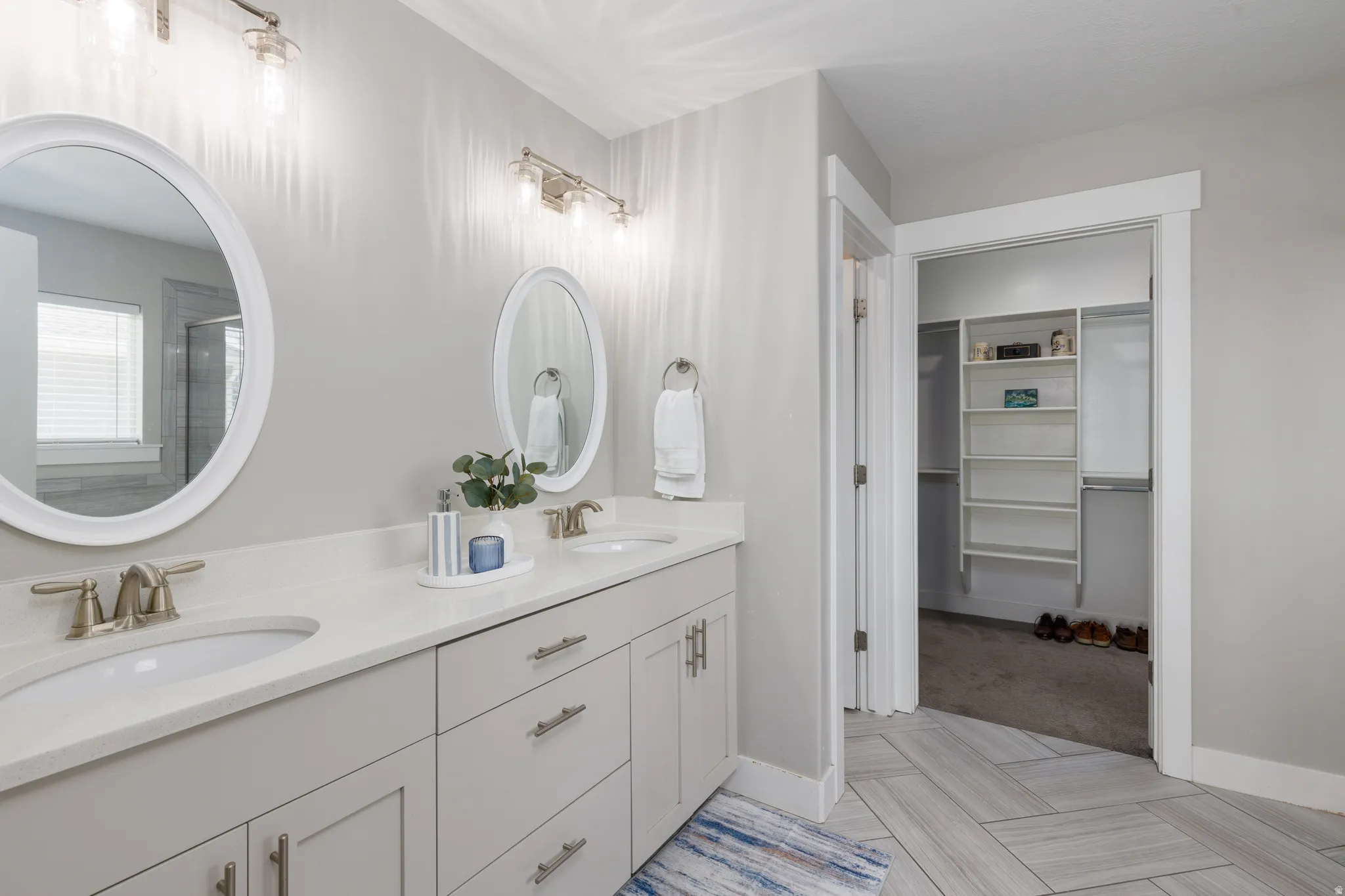 Dual oval mirrors, double vanity with light-toned cabinetry, light-toned countertops, brushed nickel hardware, and light wood-finish flooring