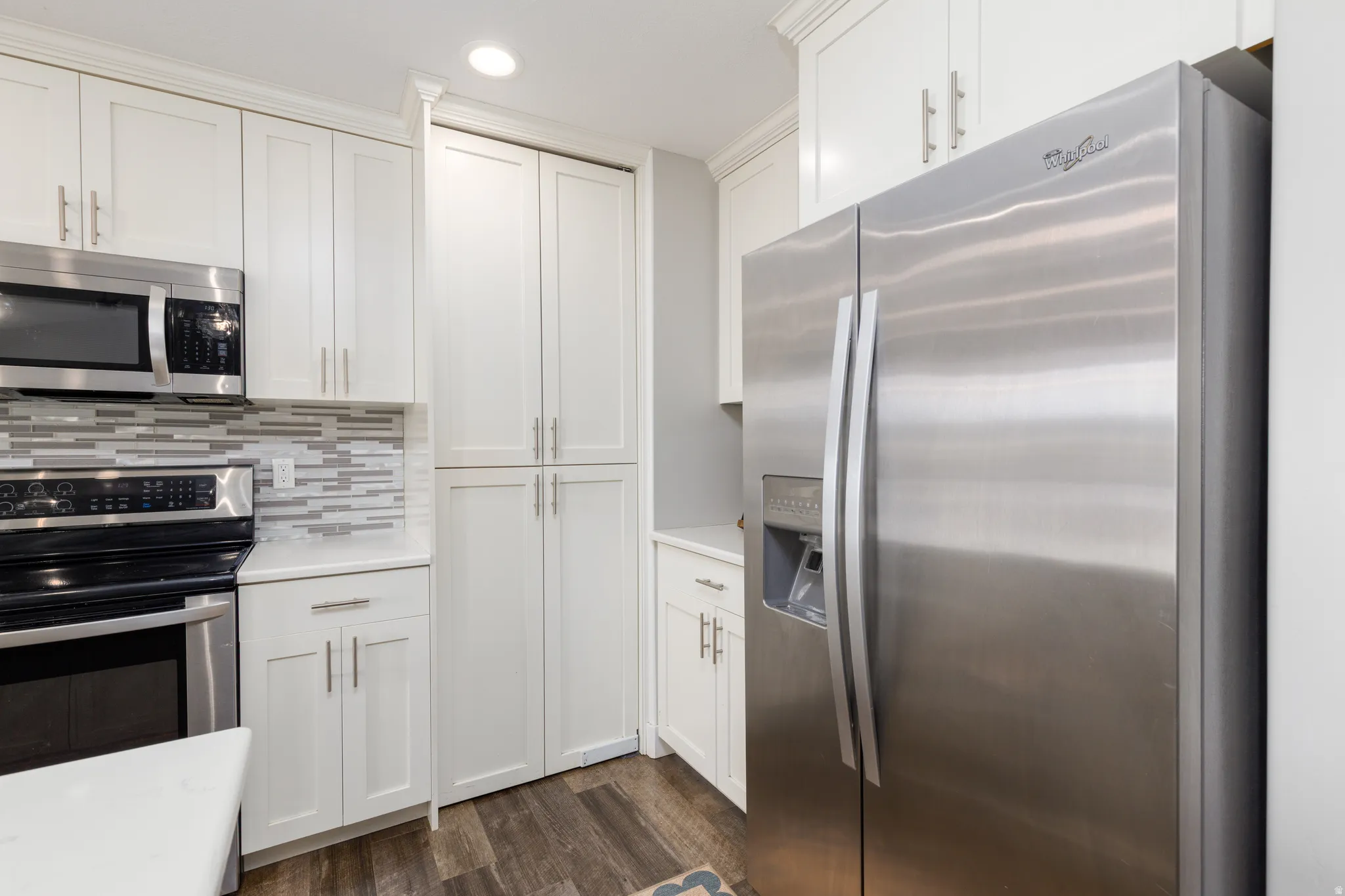 White shaker-style cabinetry with crown molding, stainless steel appliances, a mosaic tile backsplash, wood-finish flooring, and solid surface countertops