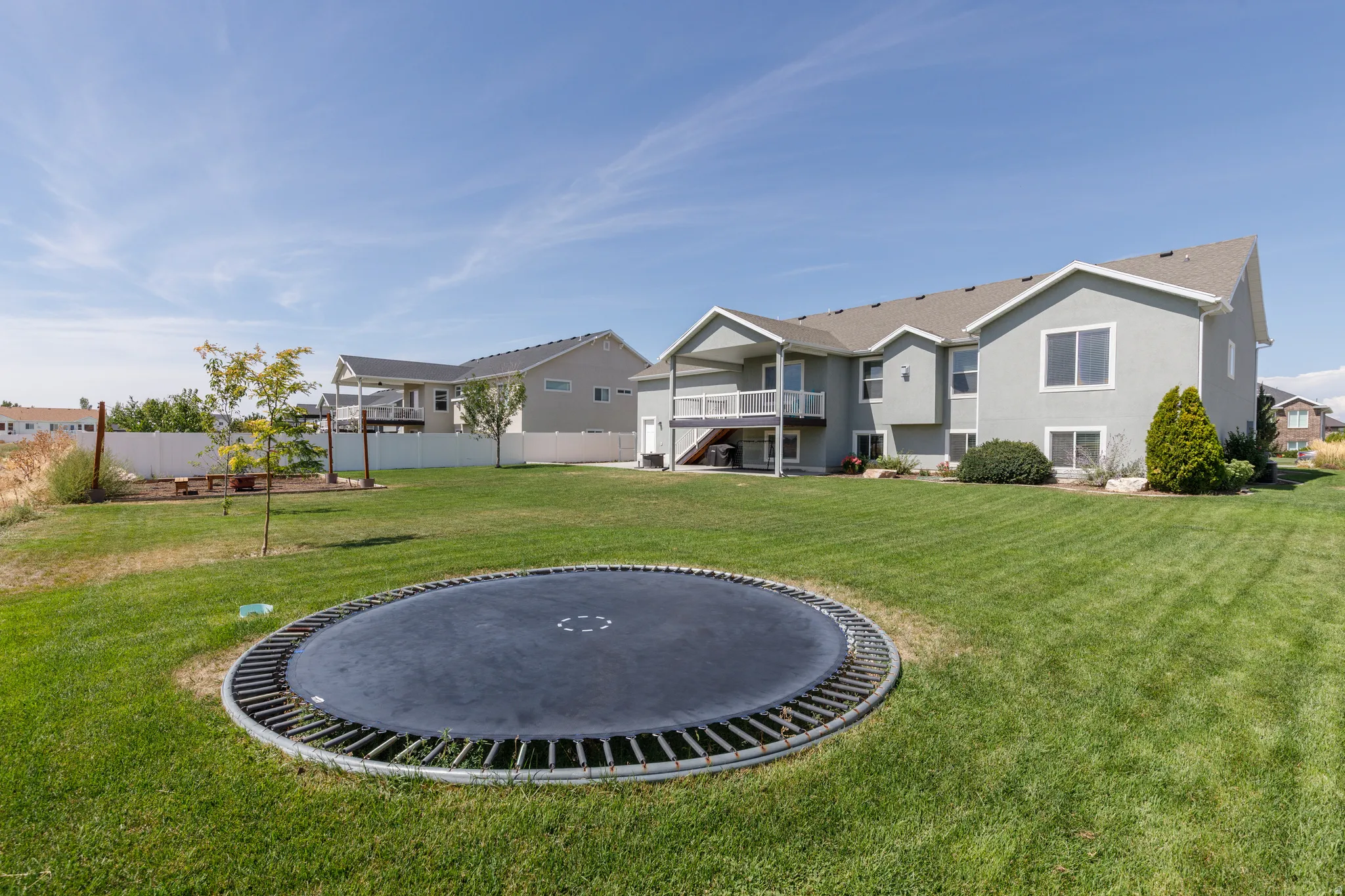 Expansive backyard featuring a large green lawn, covered deck, white vinyl fencing, and a built-in fire pit area