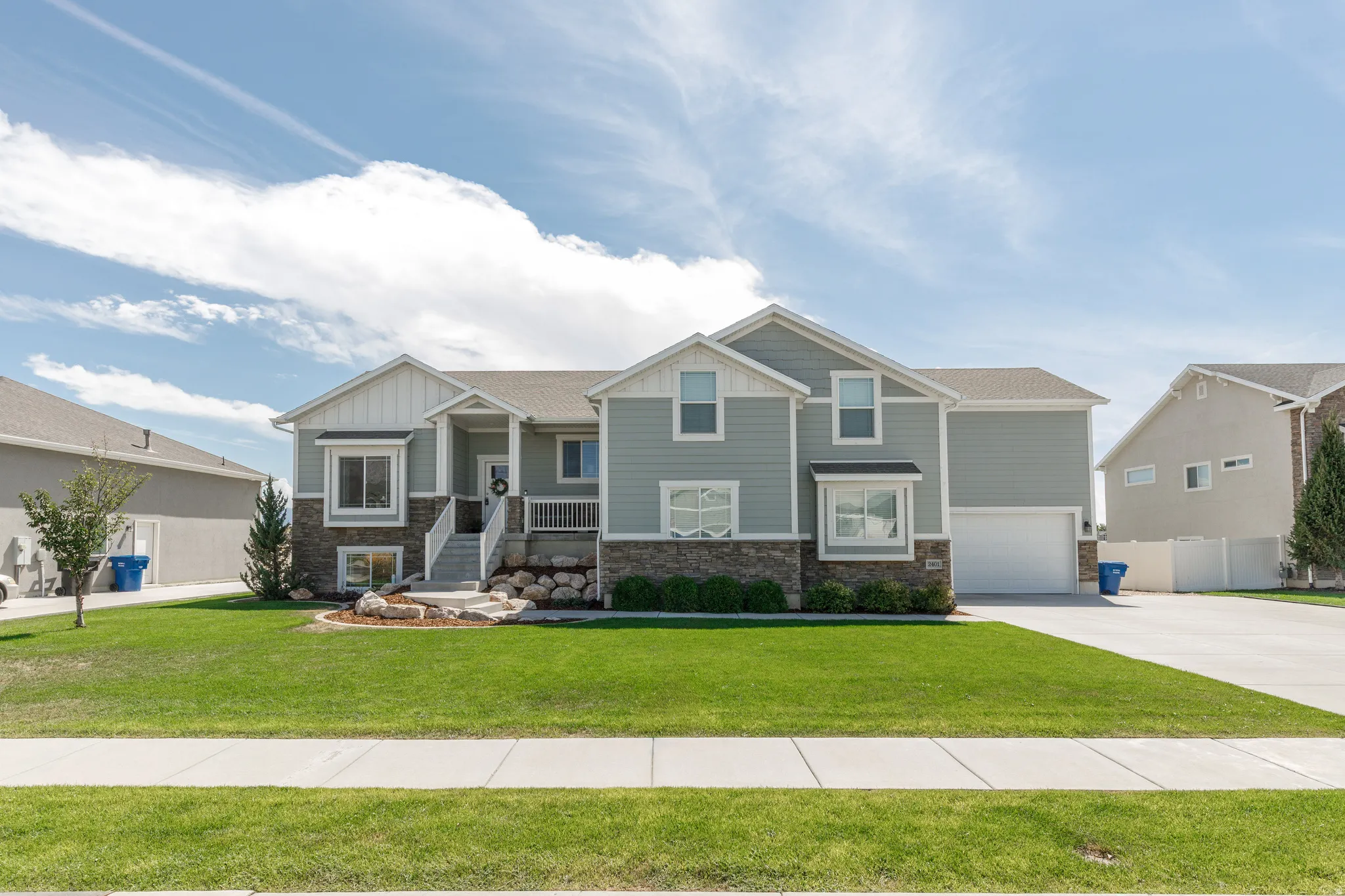 Two-story residence featuring light gray siding with white trim, a stone-clad foundation, a covered front porch, an attached garage, and a concrete driveway