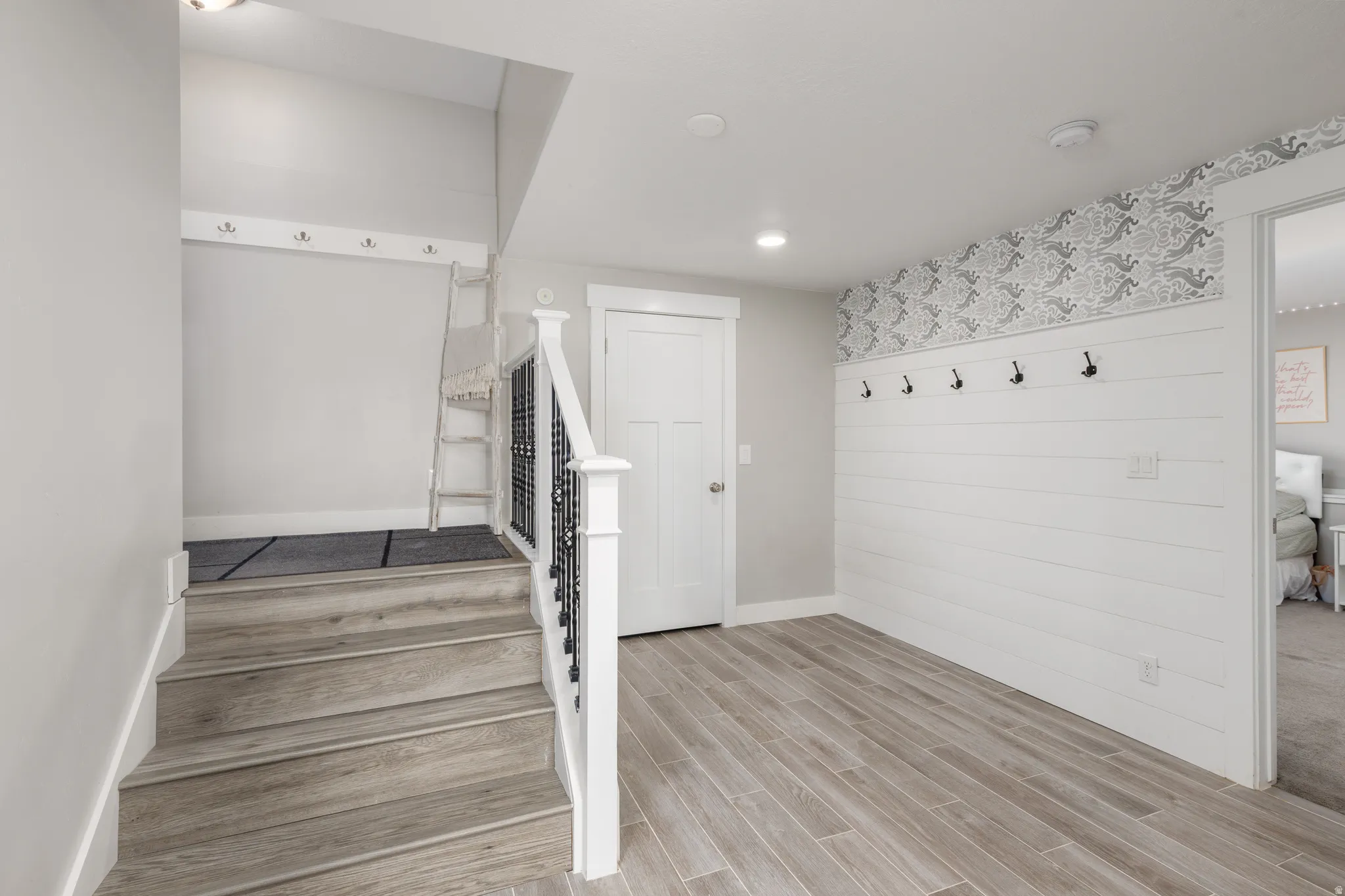 Wood-finish flooring, staircase with dark iron balusters, white shiplap accent wall, patterned wallpaper, and recessed lighting