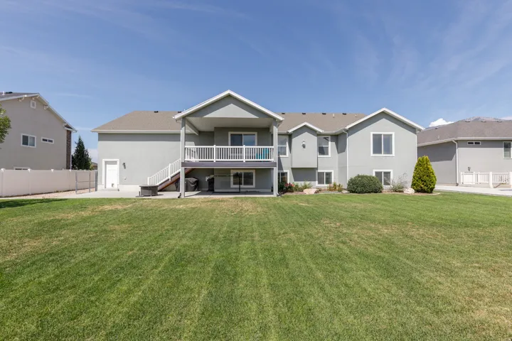 Expansive rear yard featuring a manicured lawn, stucco exterior, upper-level deck with white railings, and a concrete patio