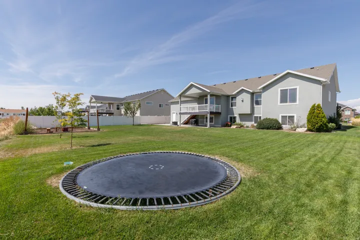 Expansive backyard featuring a large green lawn, covered deck, white vinyl fencing, and a built-in fire pit area