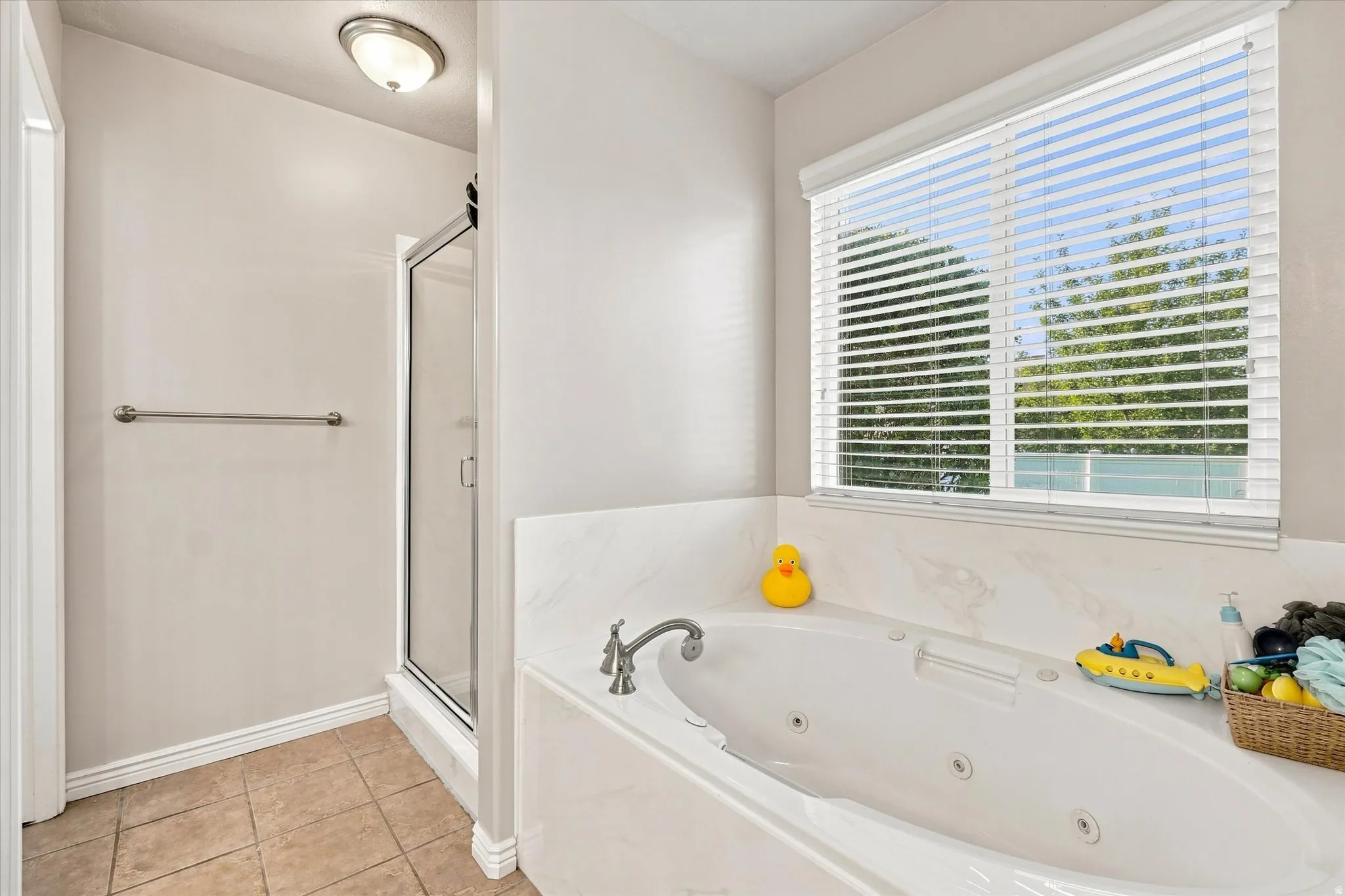 Soaking tub with integrated jets, tiled flooring, glass-enclosed shower, and a window with blinds